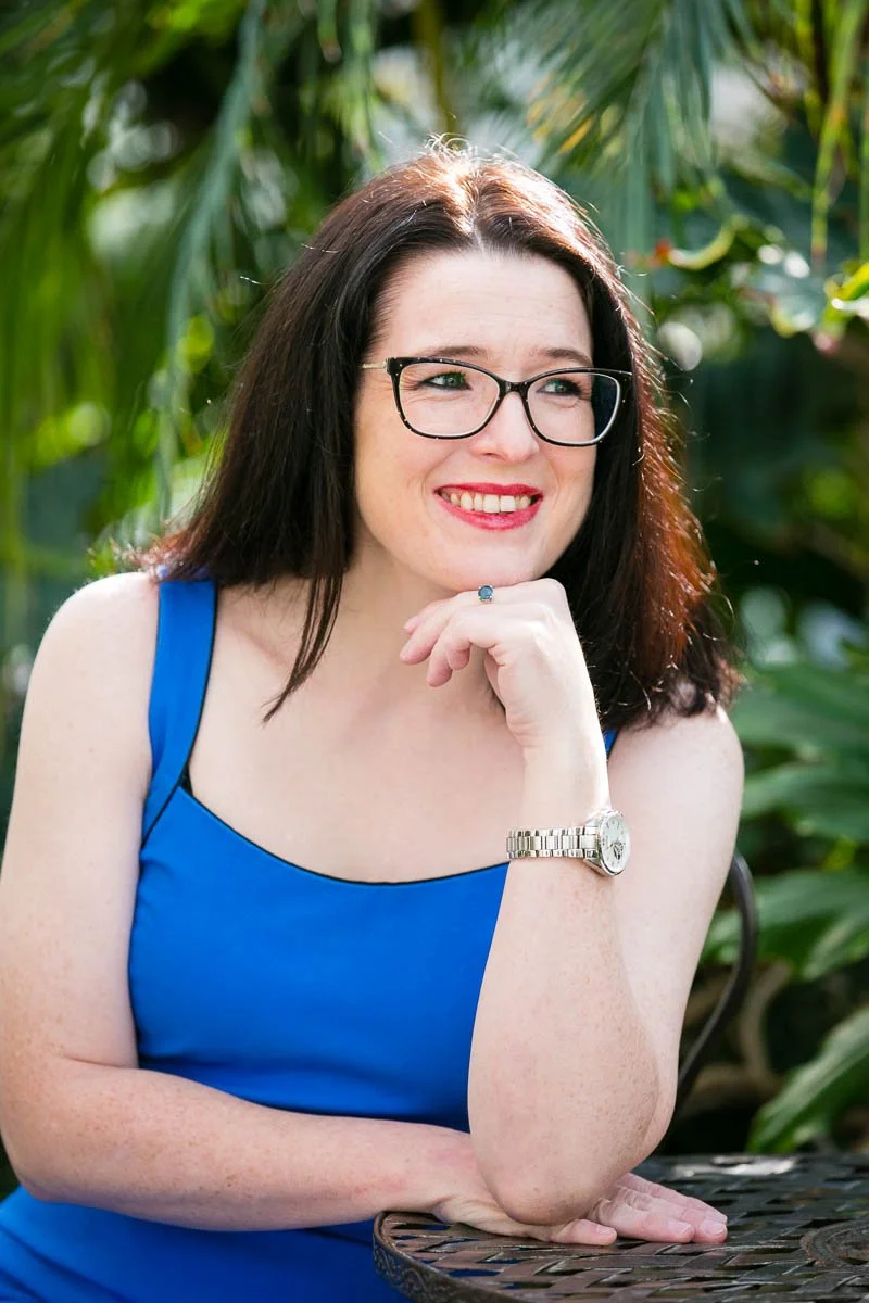 Woman with dark hair and glasses wearing a blue dress, sitting outdoors with green foliage behind her, smiling and resting her chin on her hand.