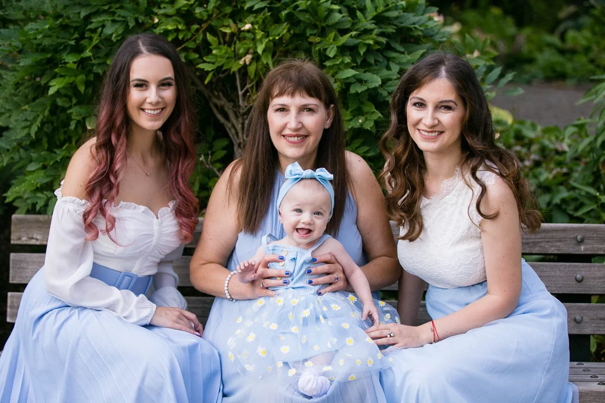 Three women and a baby girl sitting on a wooden bench outdoors with green foliage in the background, smiling at the camera. The baby girl is in the center, wearing a light blue dress with a daisy pattern and a matching blue headband.