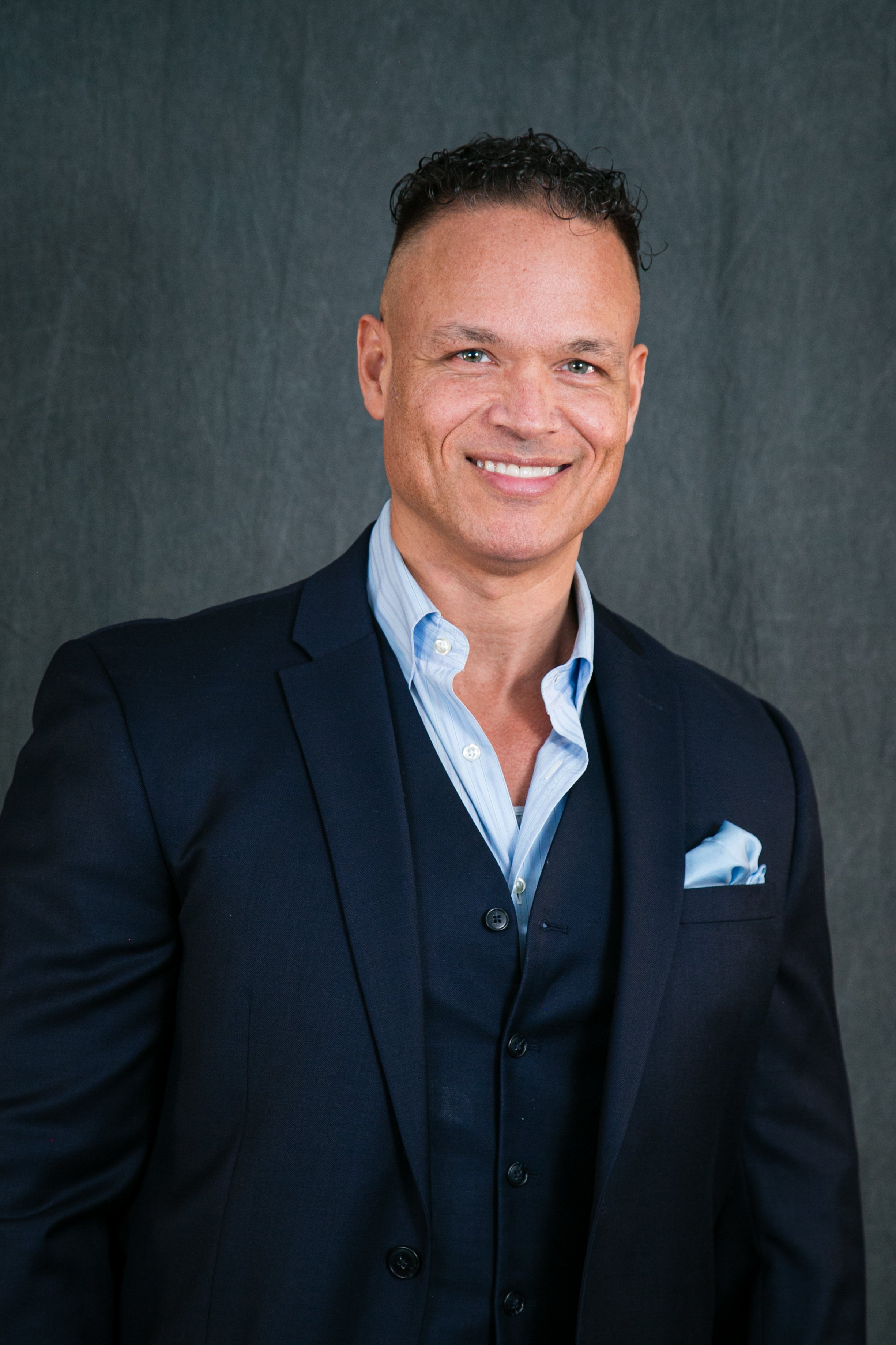 Professional man in a navy suit with light blue dress shirt, smiling, against a dark gray background.