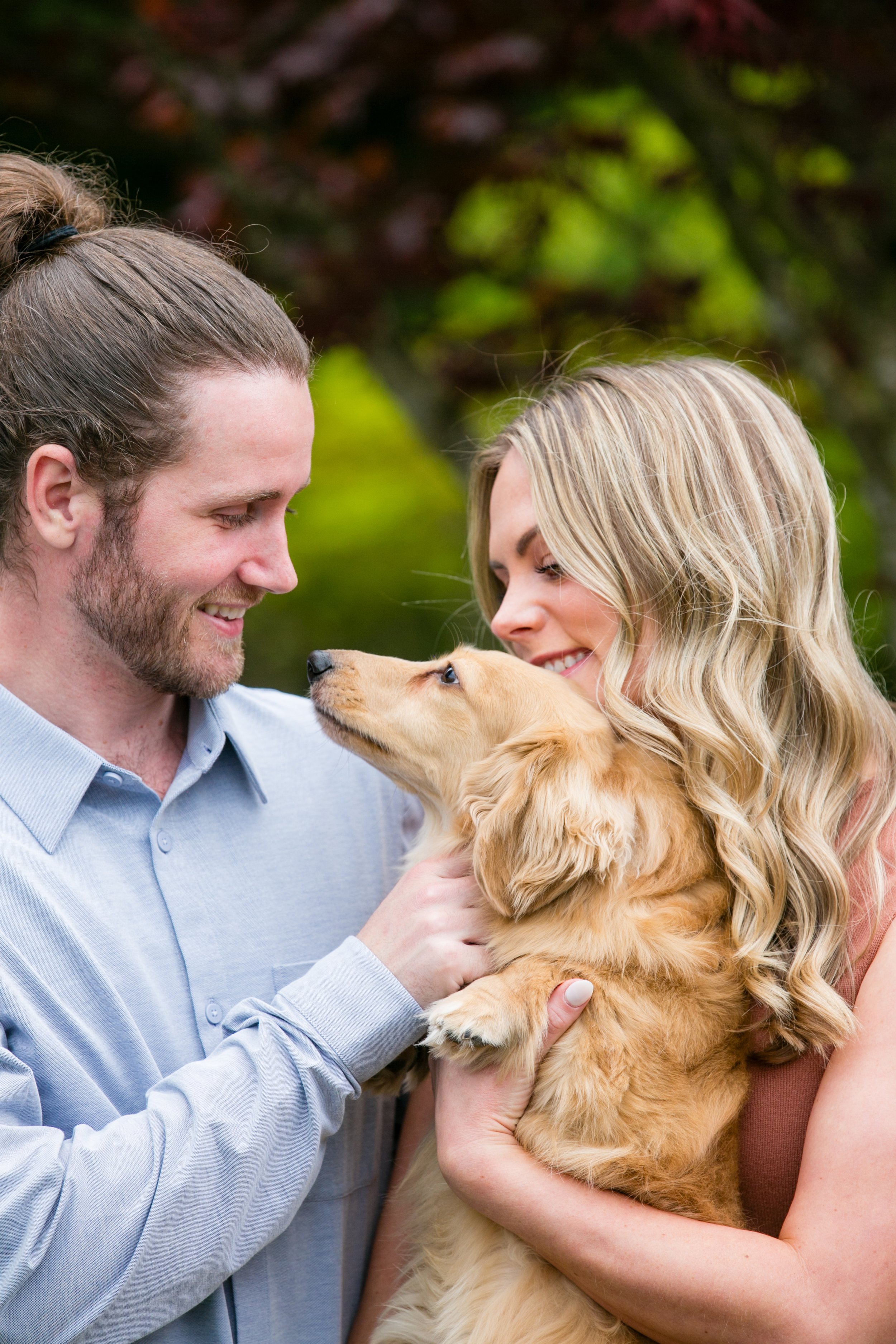 A man and woman smiling at a golden retriever puppy outdoors with a green and reddish blurry background.