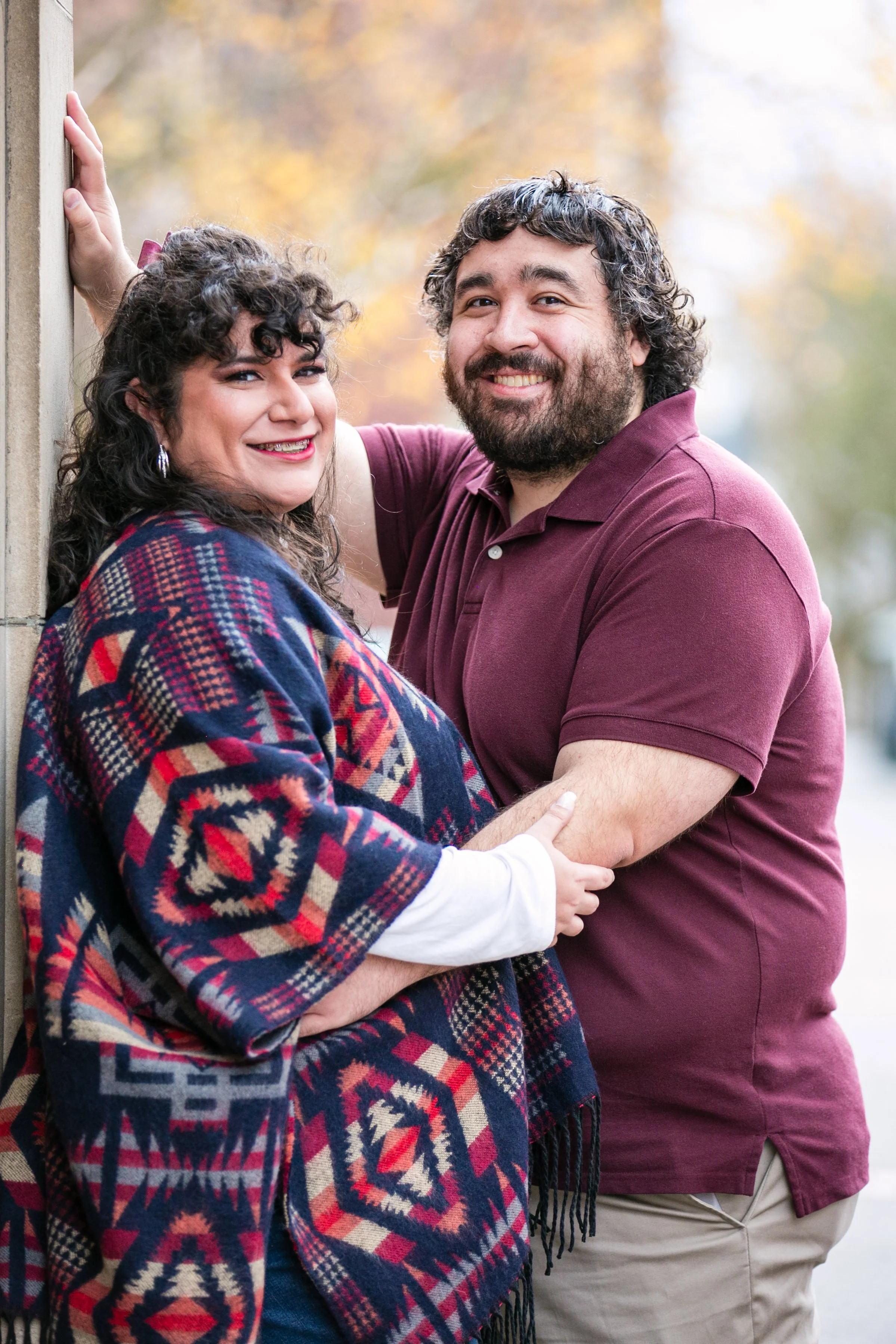 A smiling woman and man standing outdoors near a wall, with fall foliage in the background, embracing each other.