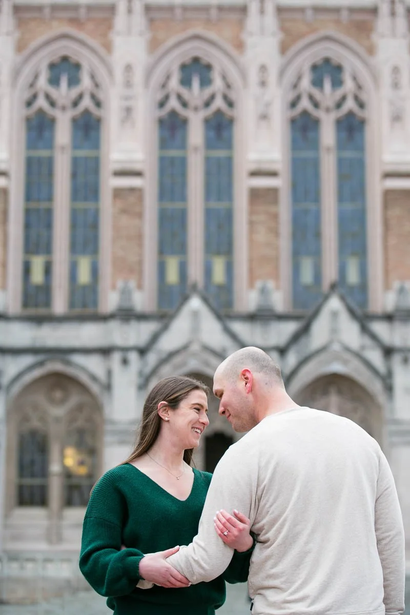 A smiling couple standing close together, outdoors, with a large Gothic-style church with tall stained glass windows in the background.
