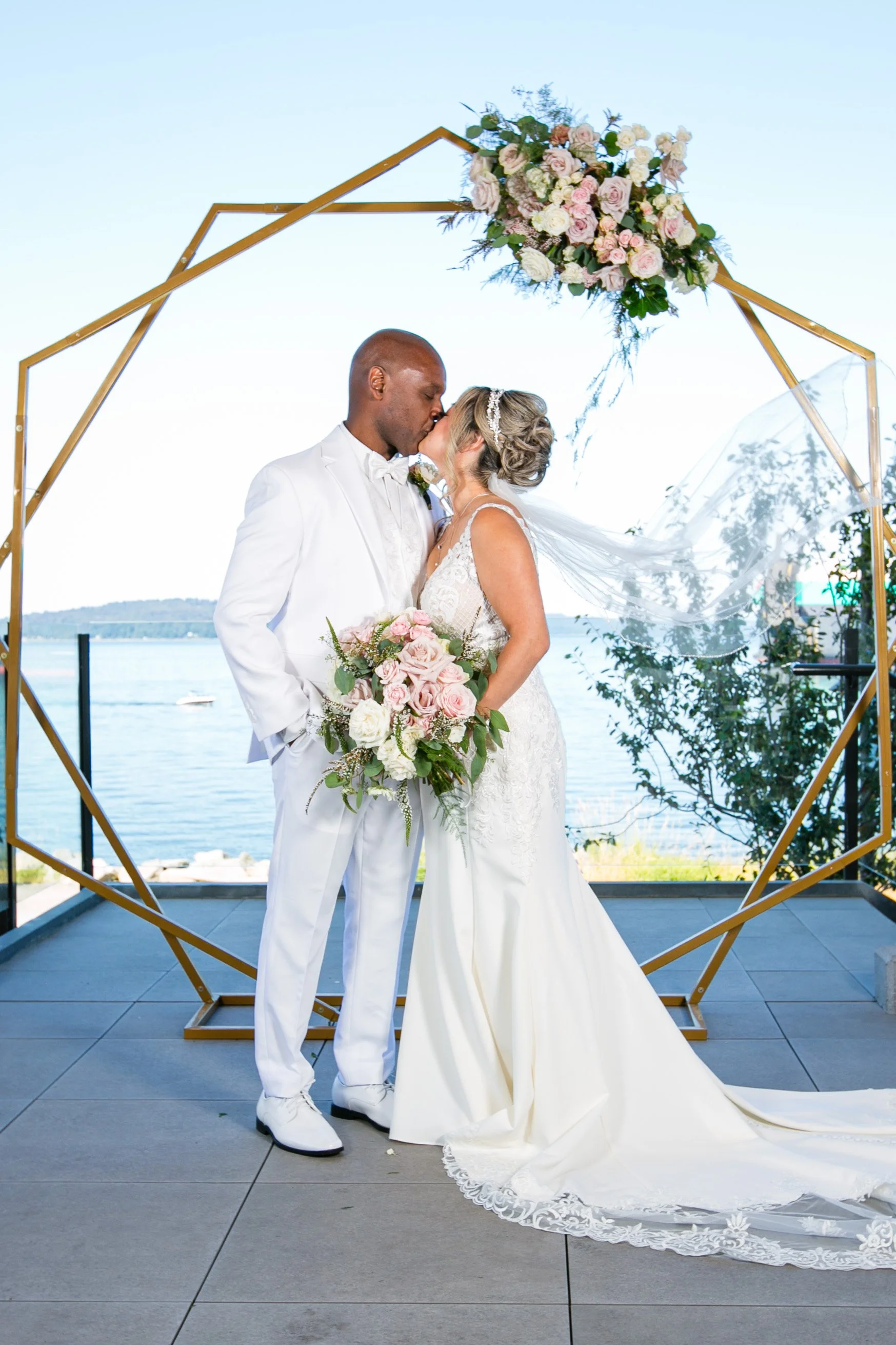 A bride and groom sharing a kiss at their wedding ceremony outdoors by the water, with a geometric gold arch decorated with pink and white flowers.
