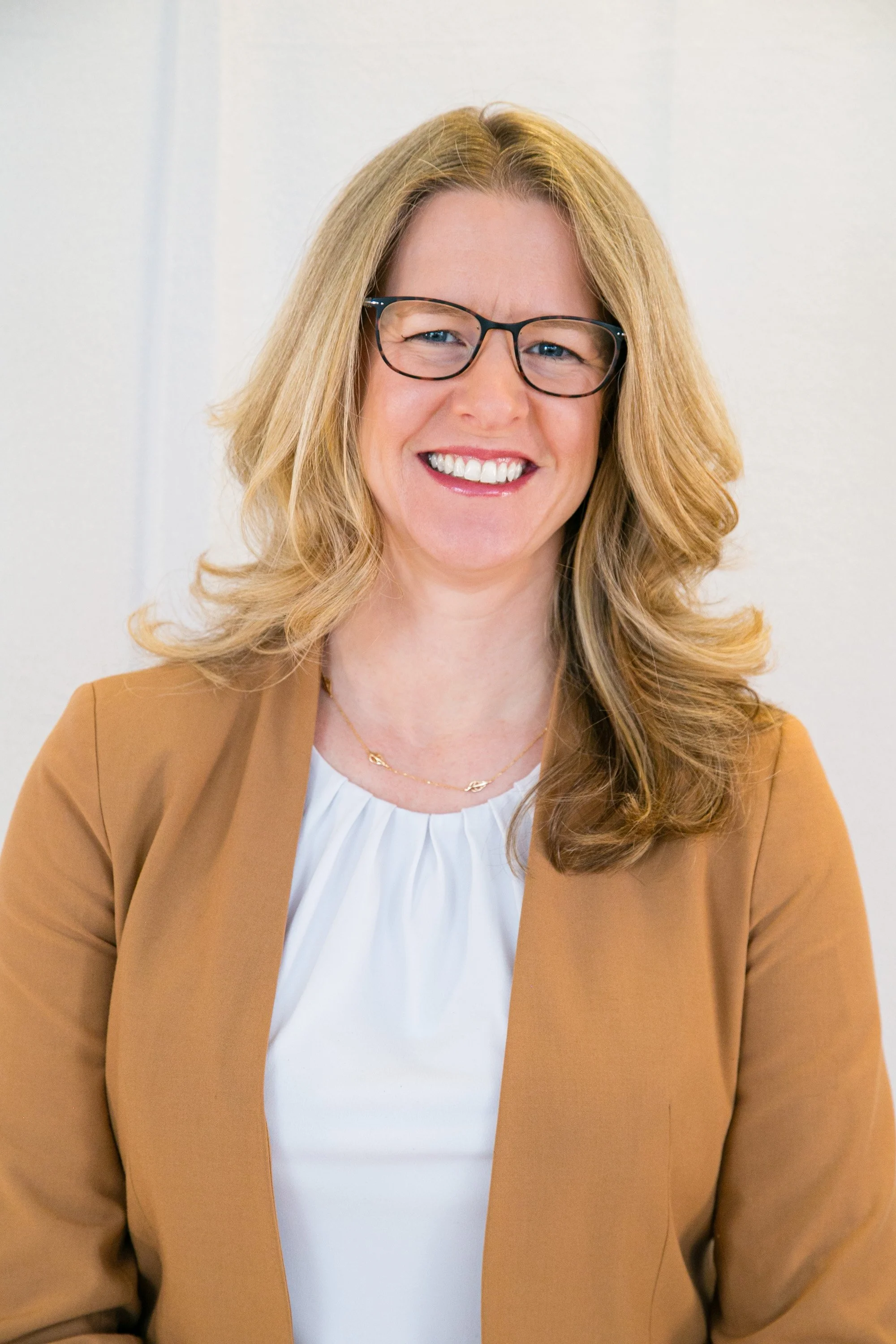 A smiling woman with blonde hair, wearing glasses, a white top, and a brown blazer, sitting indoors against a plain light-colored wall.