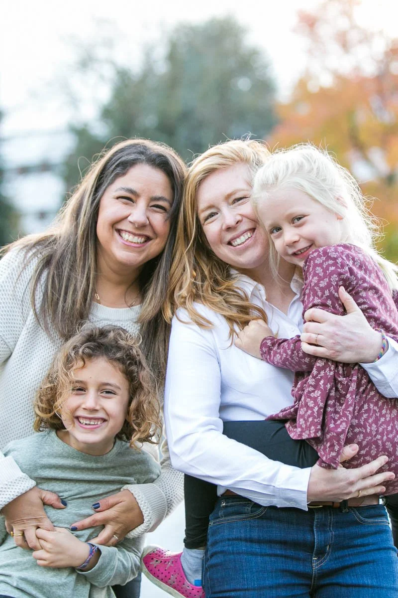 Four happy females, including a young girl, a woman with curly hair, and two women with straight hair, smiling outdoors in fall, embracing each other.