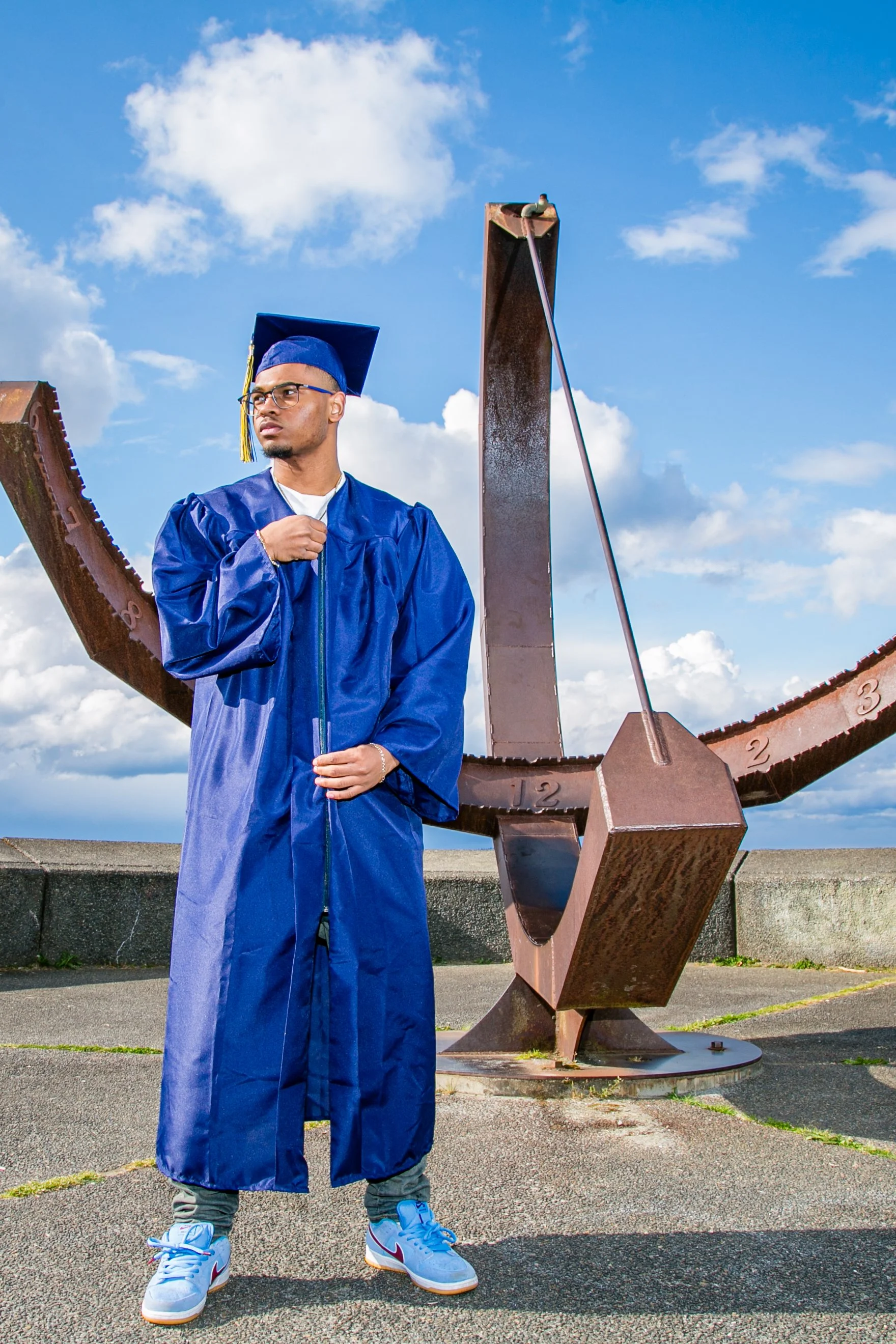 A young man in a blue graduation gown and cap standing outdoors on a concrete surface with a large rusted sundial sculpture behind him, under a partly cloudy sky.