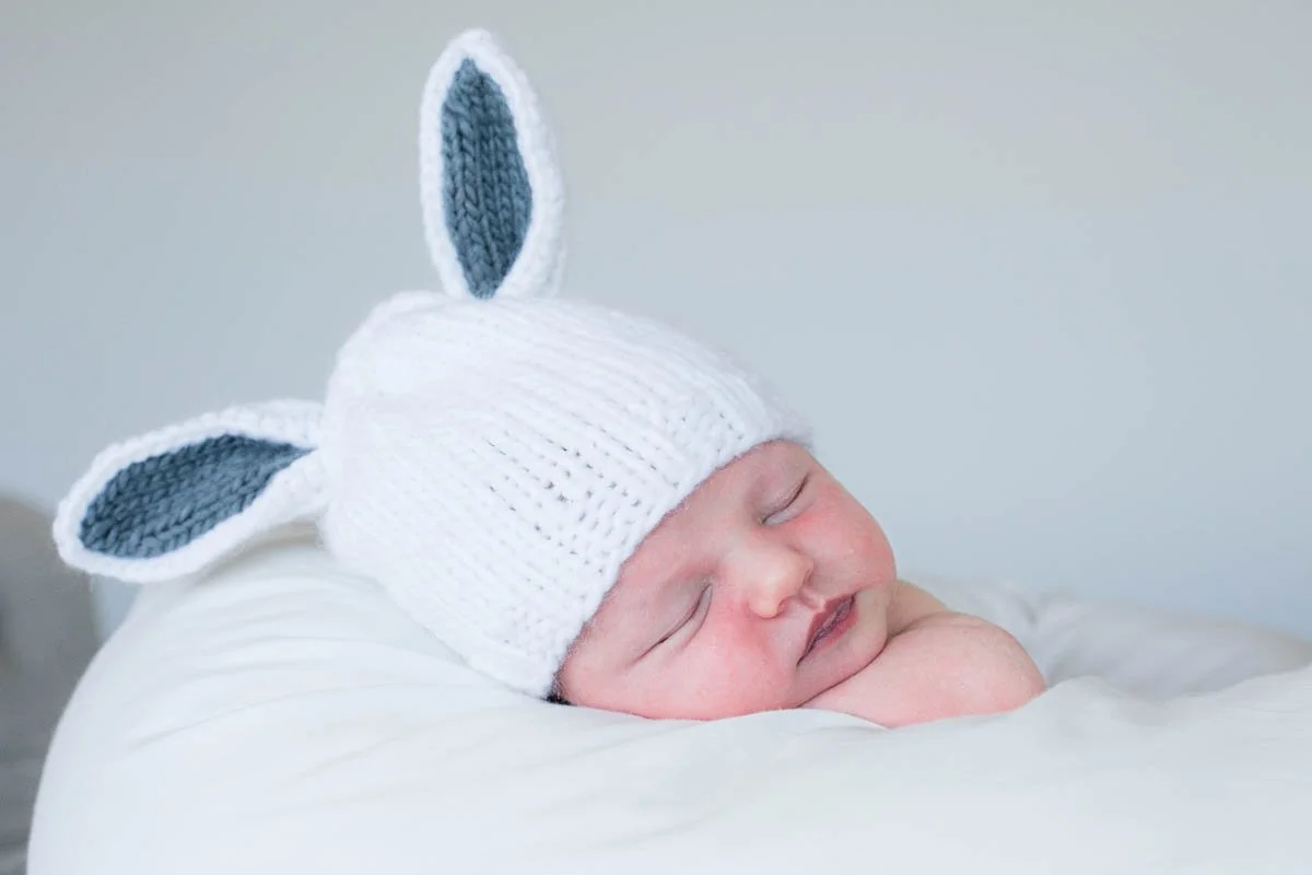 A sleeping baby wearing a white knit hat with bunny ears, lying on a white surface.