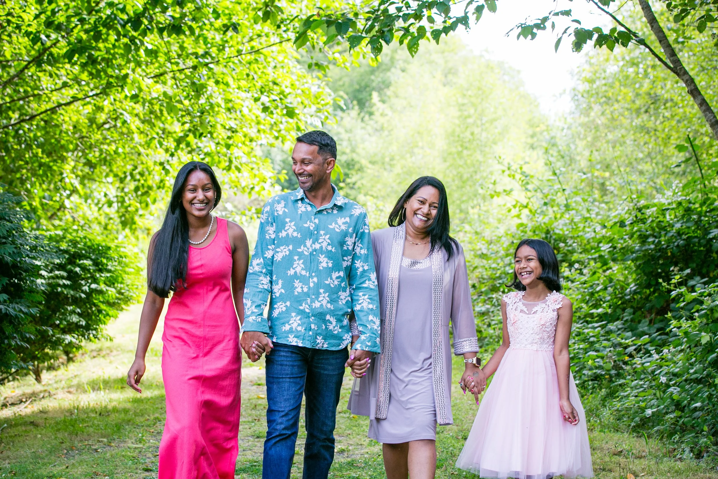 A family of four, two adults and two children, walking hand in hand outdoors in a lush green park, smiling and enjoying a sunny day.