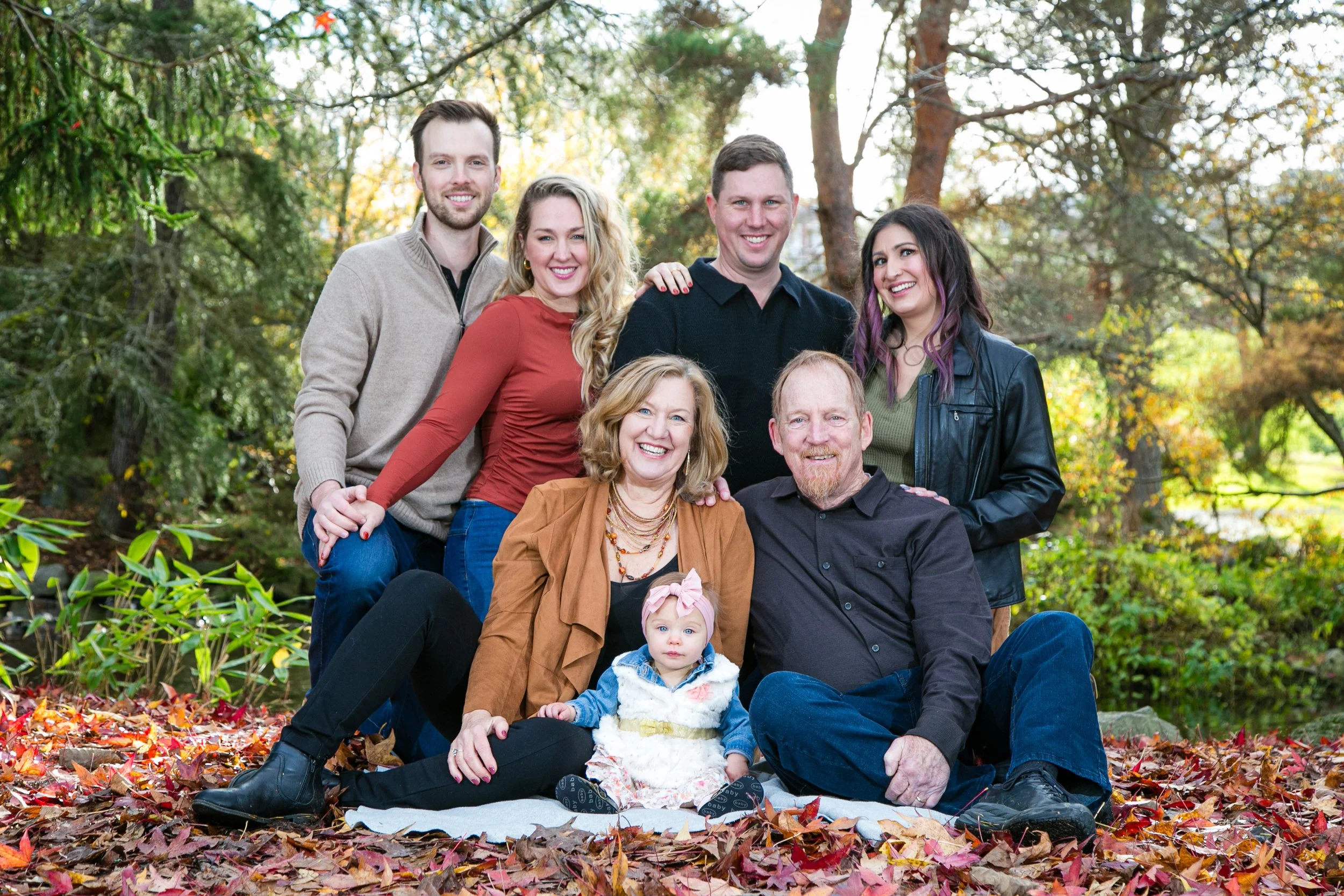 Family group photo outdoors during fall, with leaves on the ground and trees in the background. Six adults and a baby sitting or standing close together, smiling at the camera.