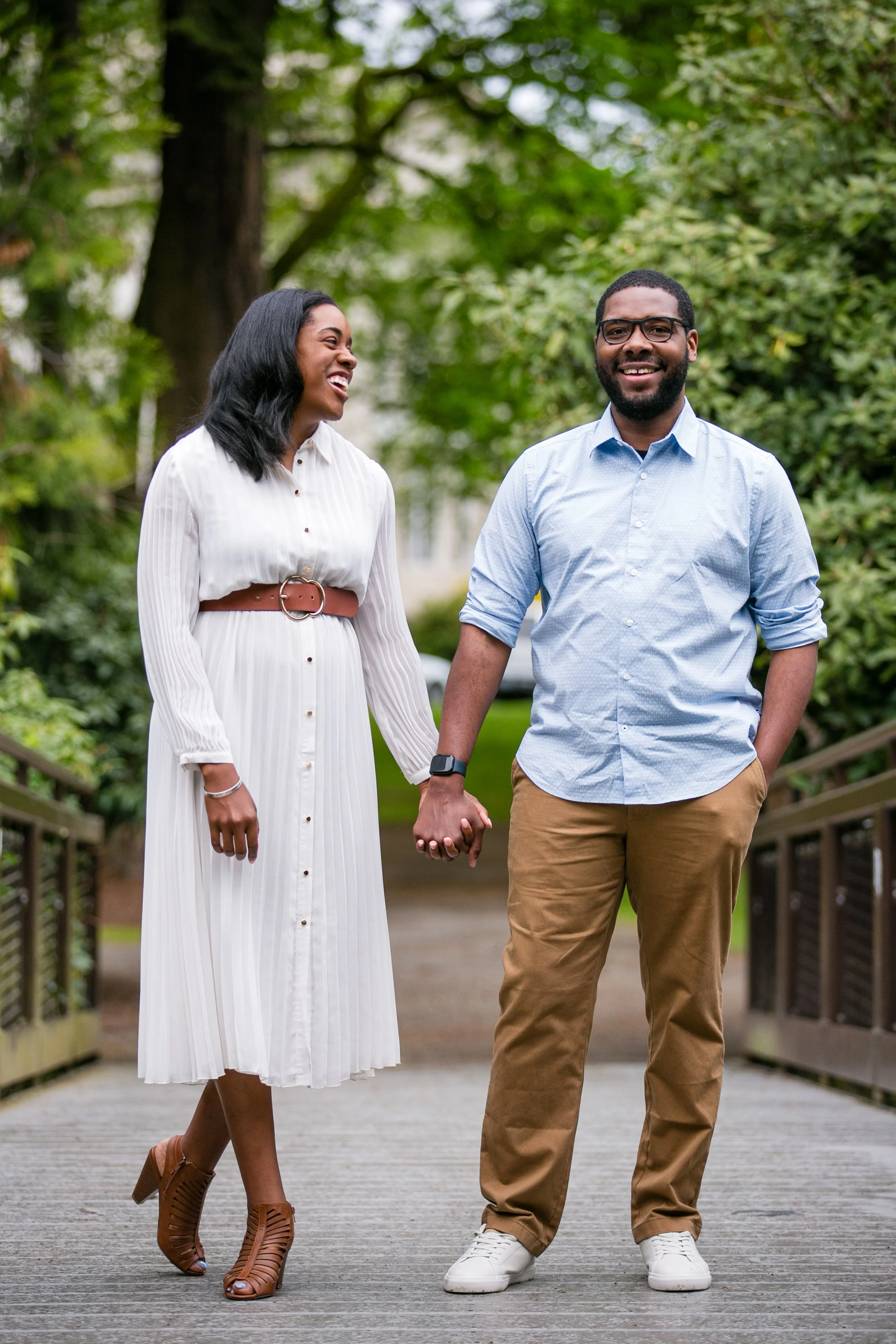 A couple walking hand-in-hand on a park bridge, smiling and enjoying each other's company surrounded by lush green trees.