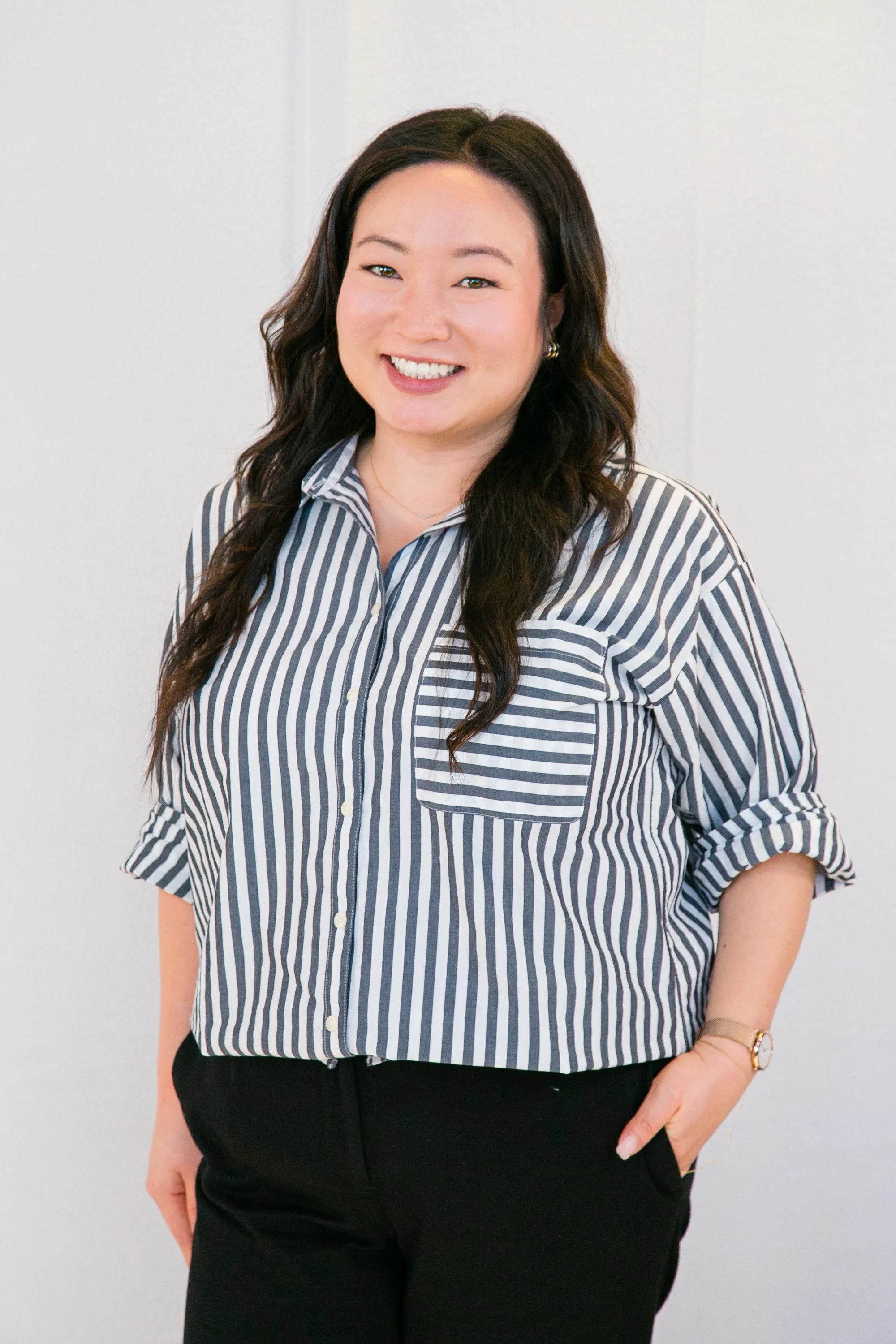 A smiling woman with long wavy dark hair, wearing a blue and white striped button-up shirt and black pants, standing against a plain white wall.