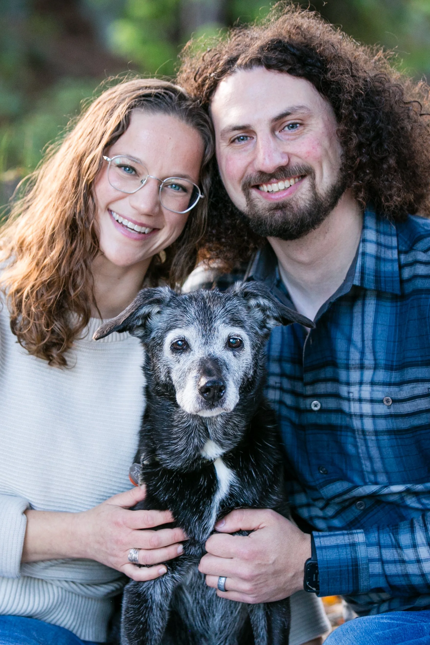 A smiling couple with glasses and curly hair, holding a gray and black dog, outdoors surrounded by trees.