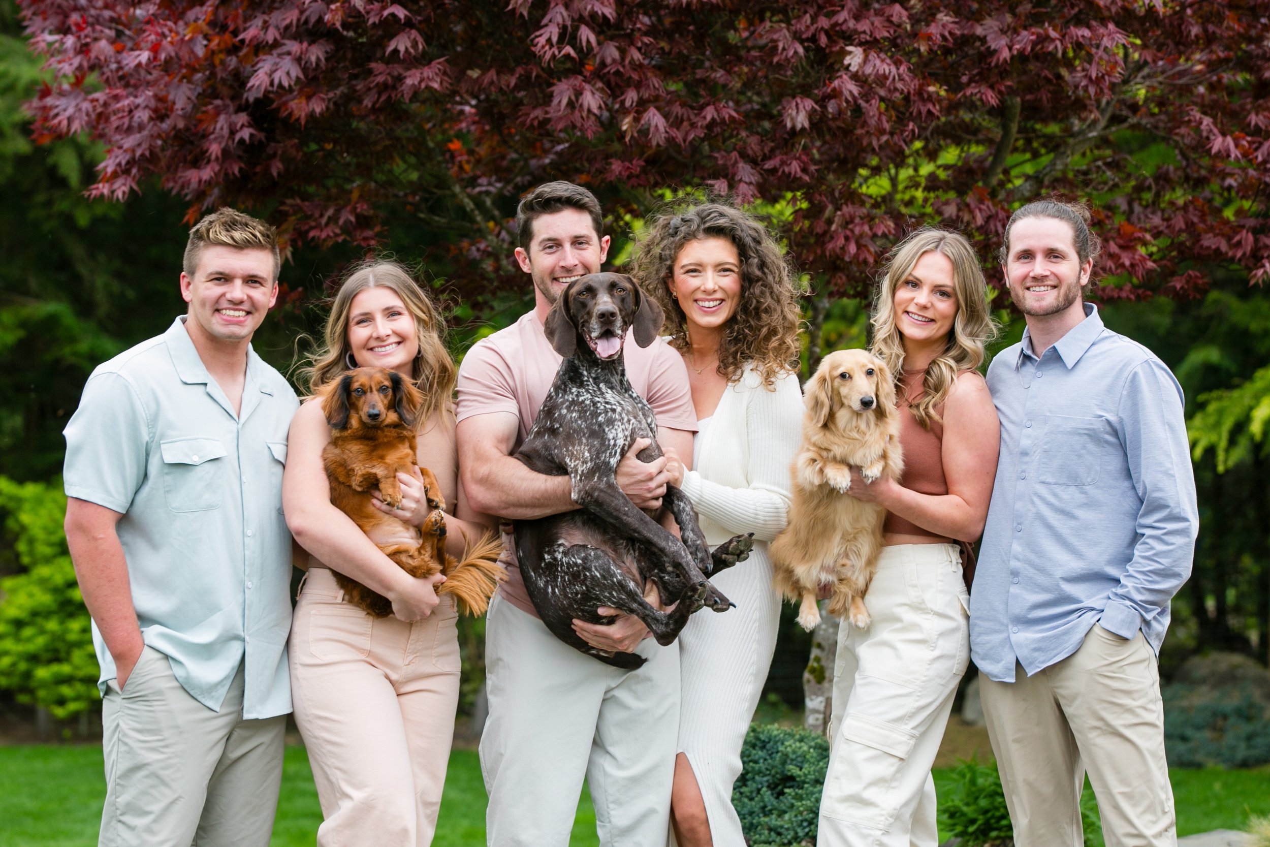 Group of seven people smiling outdoors, each holding a dog, with trees and greenery in the background.