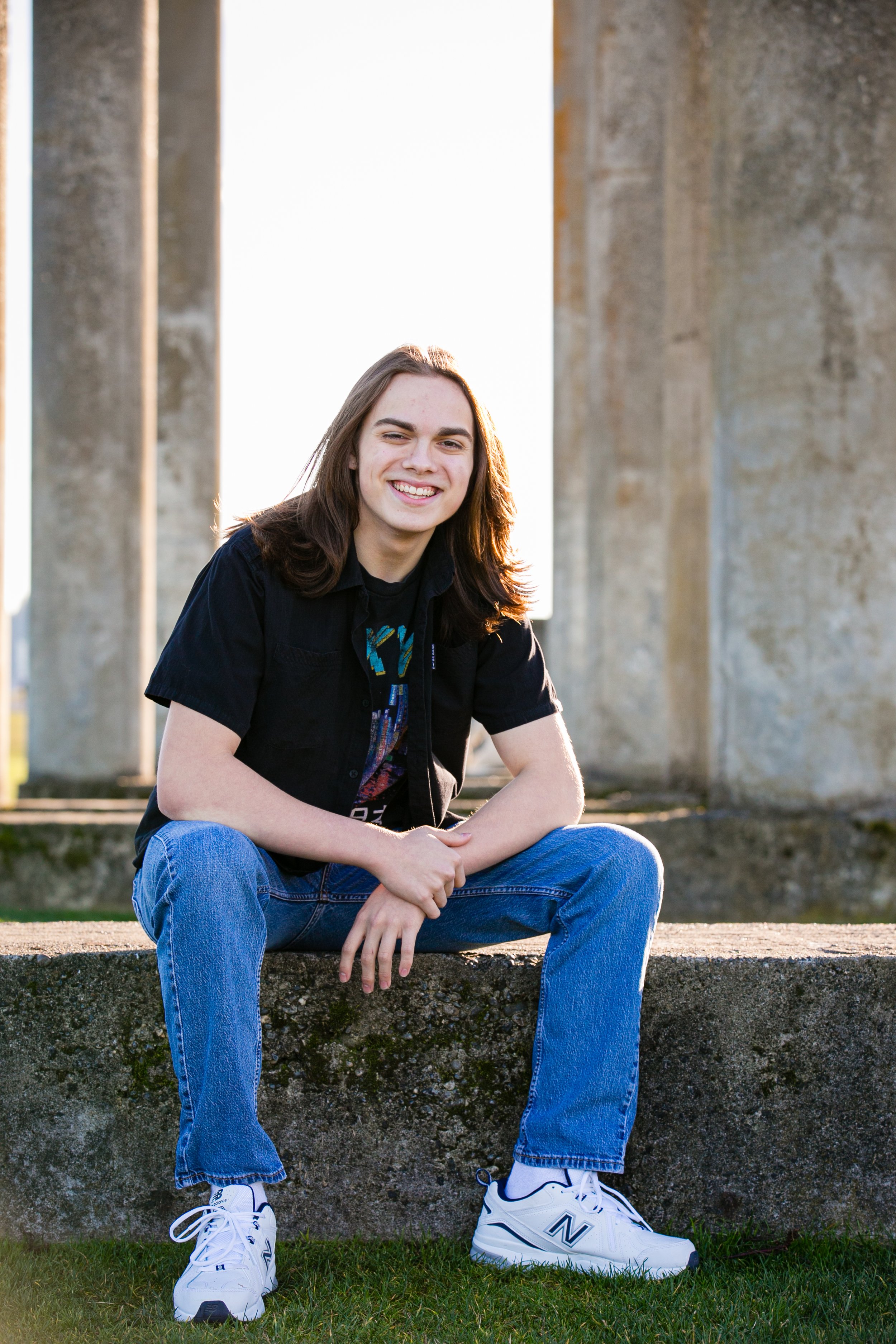 A young man with long brown hair sitting on a concrete ledge under large concrete pillars with a bright sky behind him, smiling at the camera.