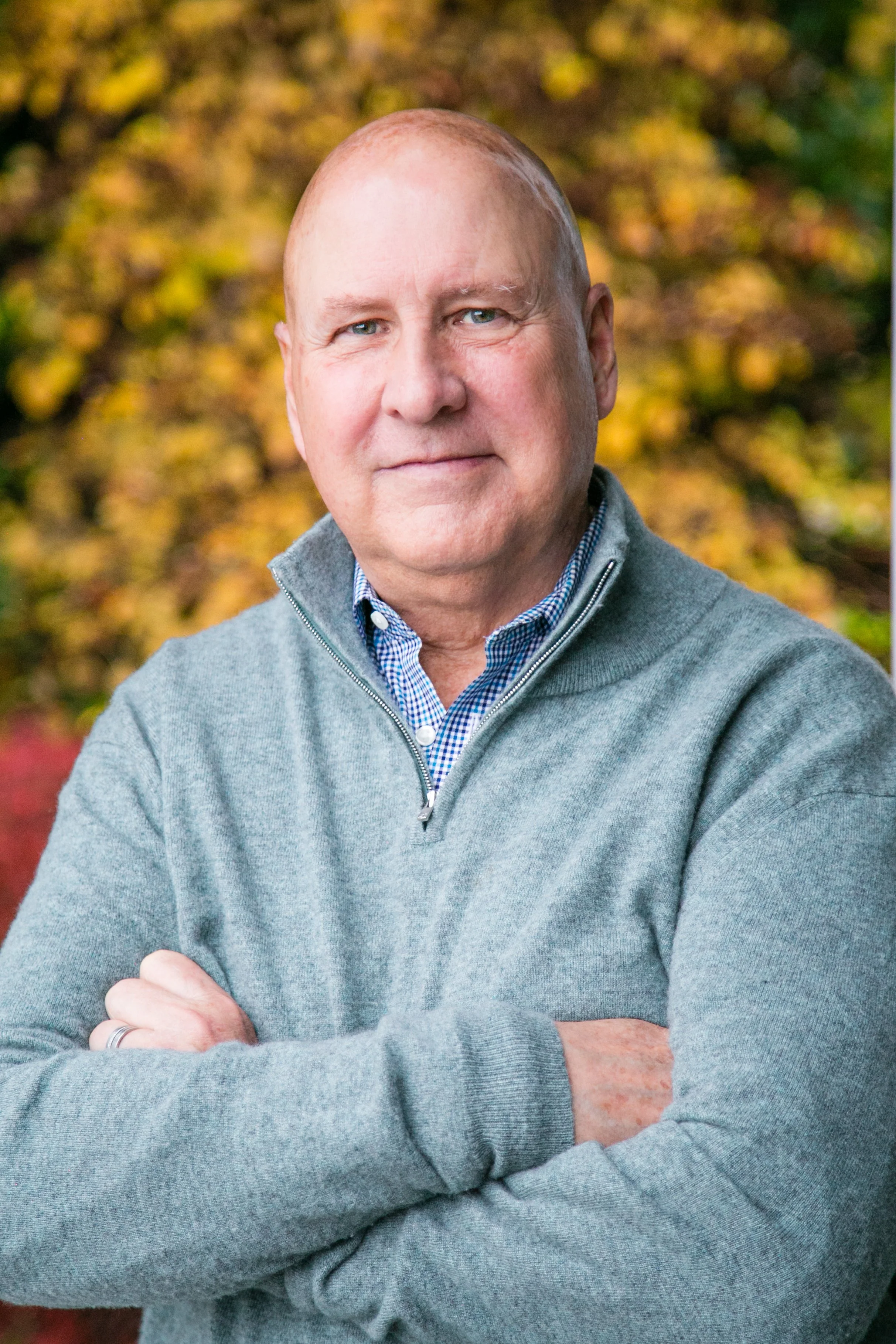 A middle-aged man with a bald head, blue eyes, and a fair complexion standing outdoors with arms crossed. He is wearing a gray quarter-zip sweater over a blue checkered shirt, with autumn-colored trees in the background.