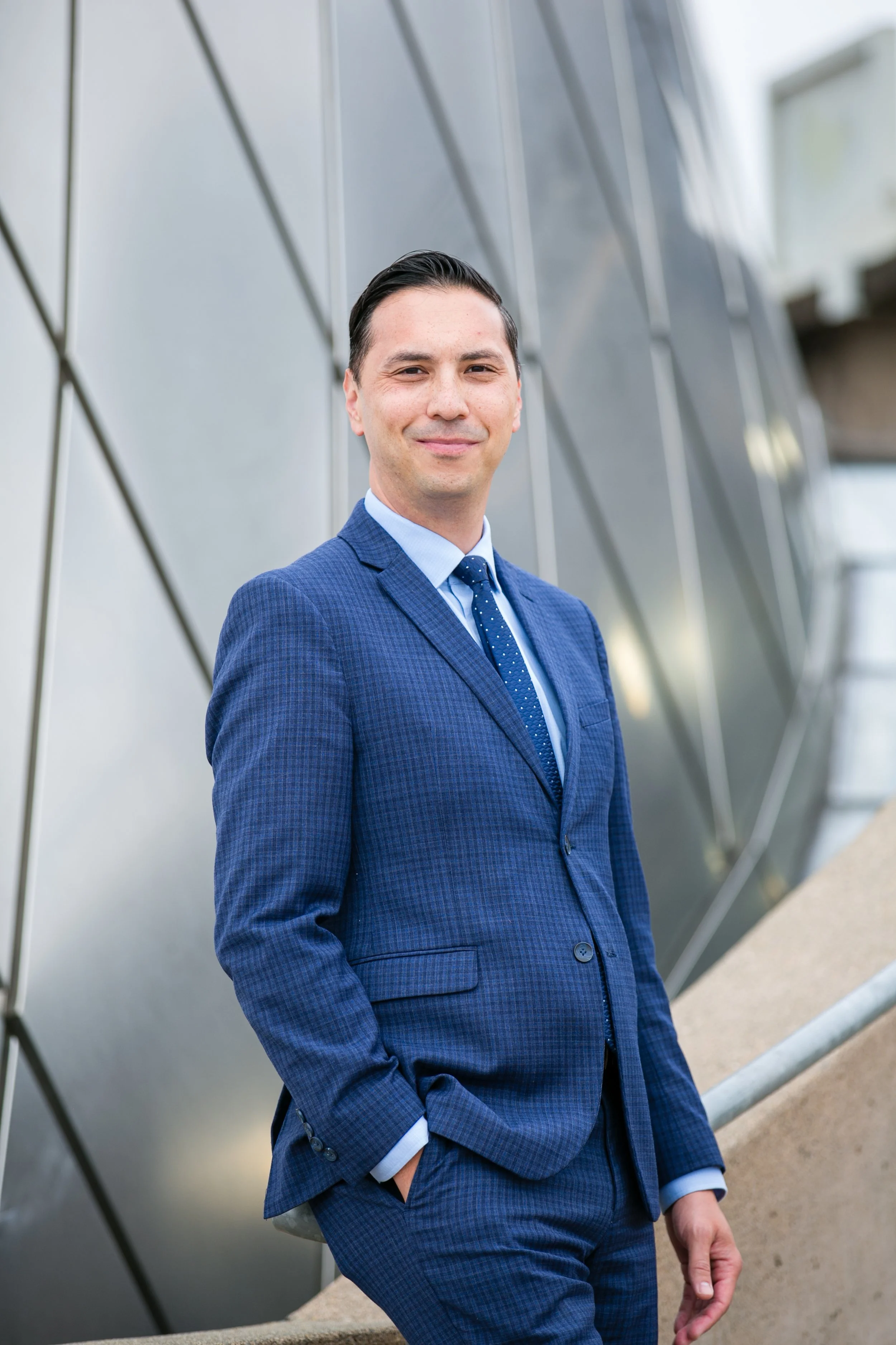 A man in a blue checkered suit, light blue shirt, and dark blue tie with white polka dots, standing outdoors in front of a modern metallic building with angular panels, smiling at the camera with one hand in his pocket.