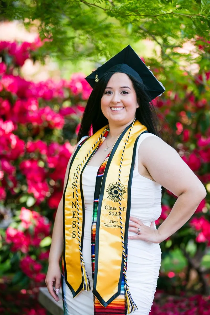 A young woman wearing a black graduation cap and gown with a yellow Latin honors stole and multicolored cords, smiling outdoors with vibrant pink flowers and green foliage in the background.