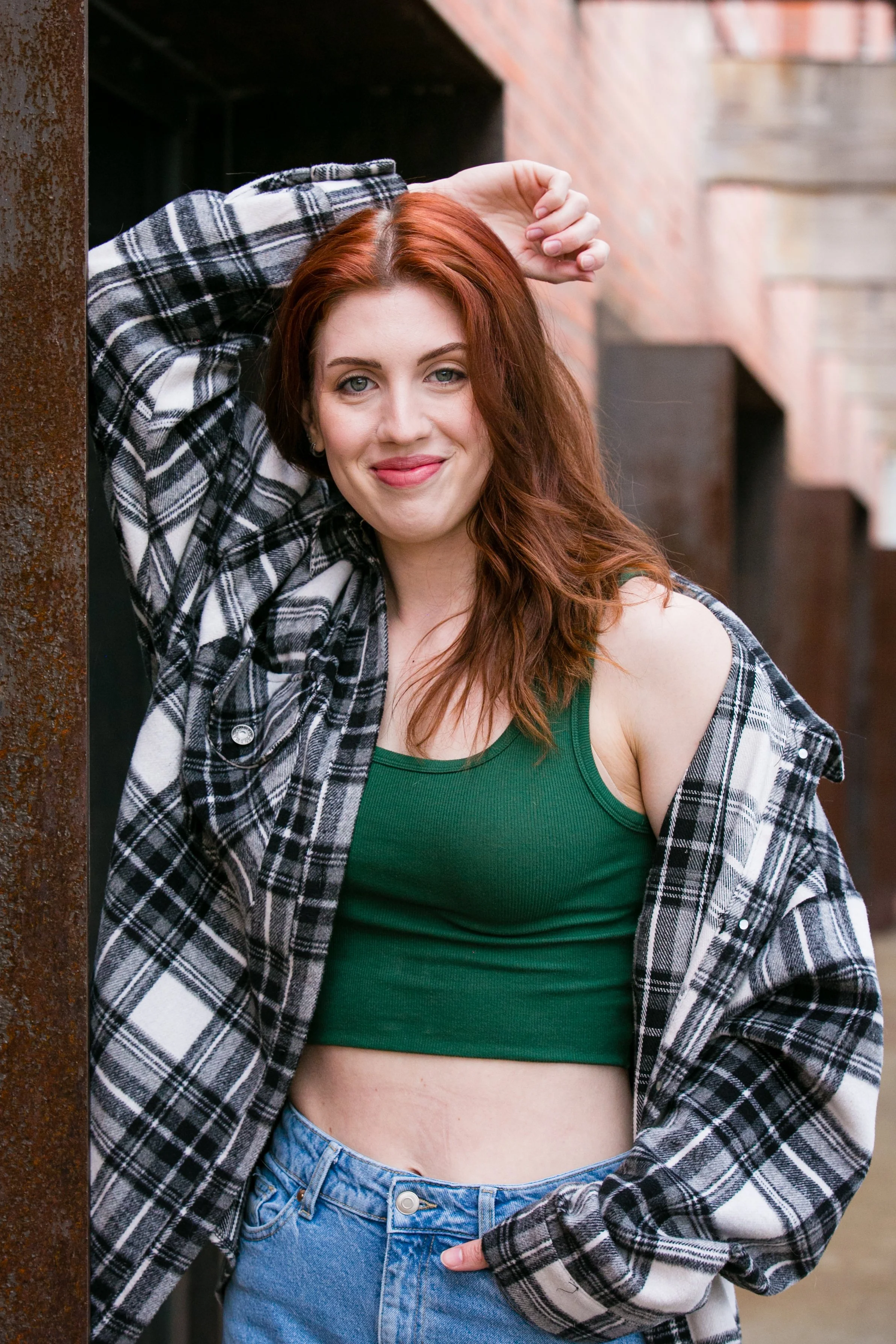 A young woman with red hair, wearing a green tank top, a plaid shirt, and jeans, leaning against a metal pillar outdoors.