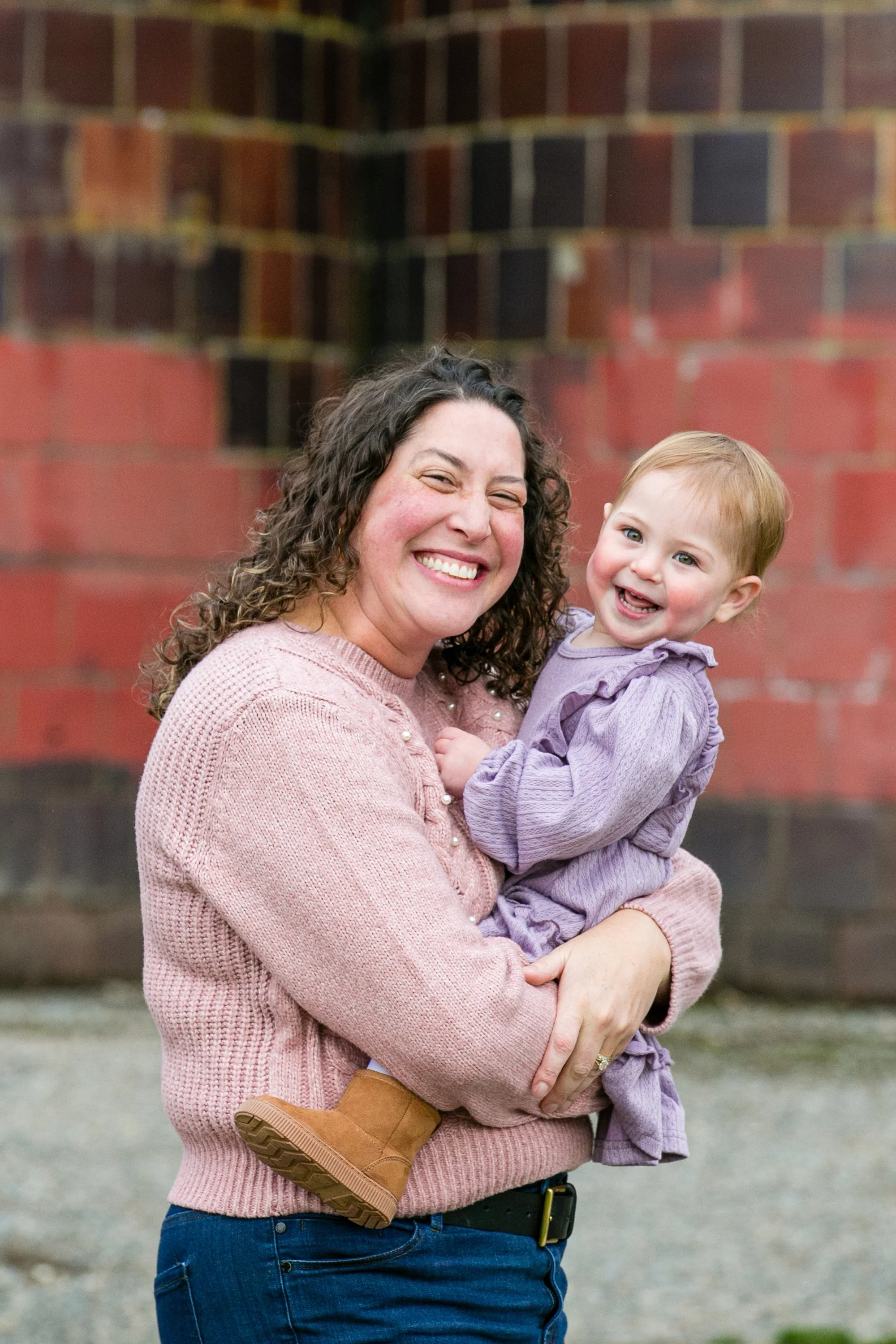 A woman with curly hair holding a young girl with blonde hair, both smiling, standing outdoors near a brick wall.