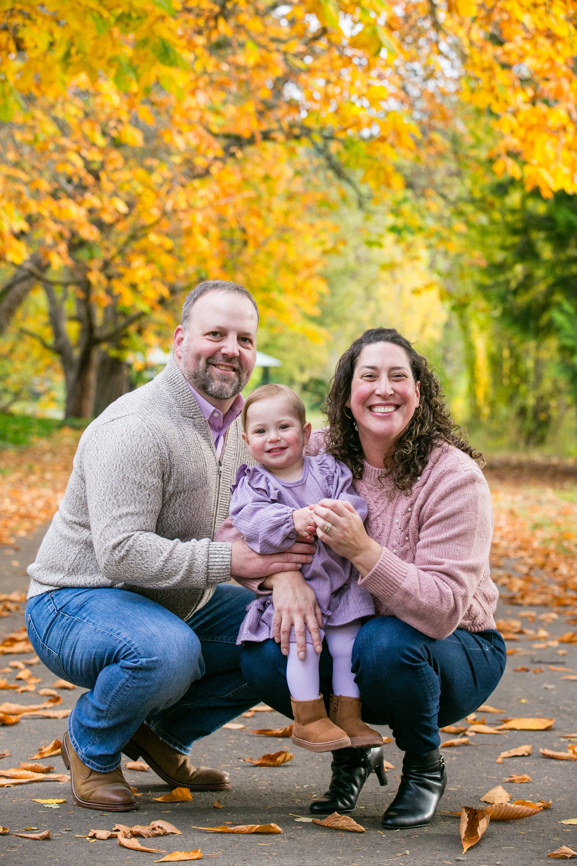 A family of three posing outdoors in a park during autumn, with colorful fall leaves on trees and scattered on the ground, smiling at the camera.