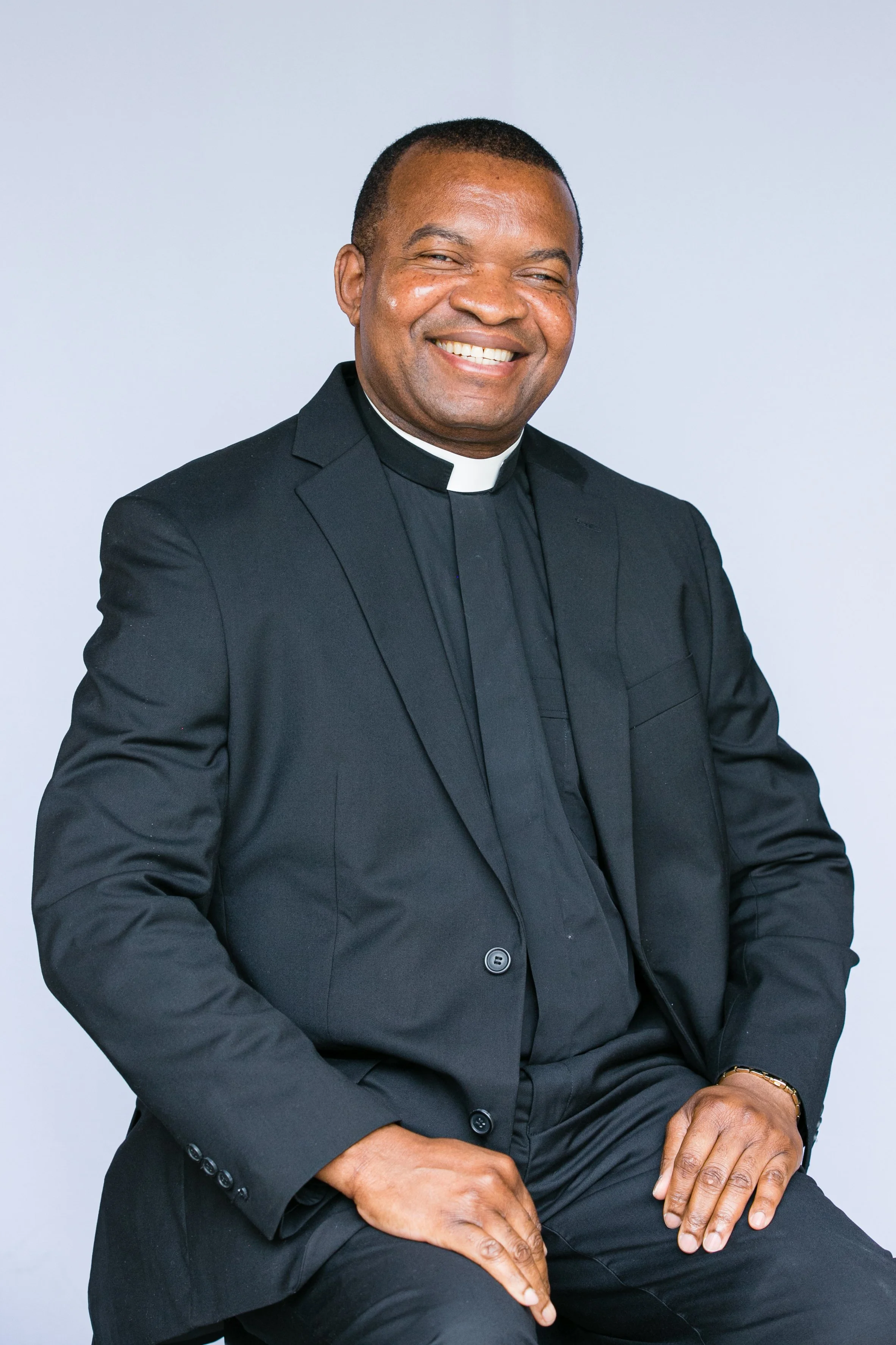 A smiling man dressed as a Catholic priest poses against a light gray background.