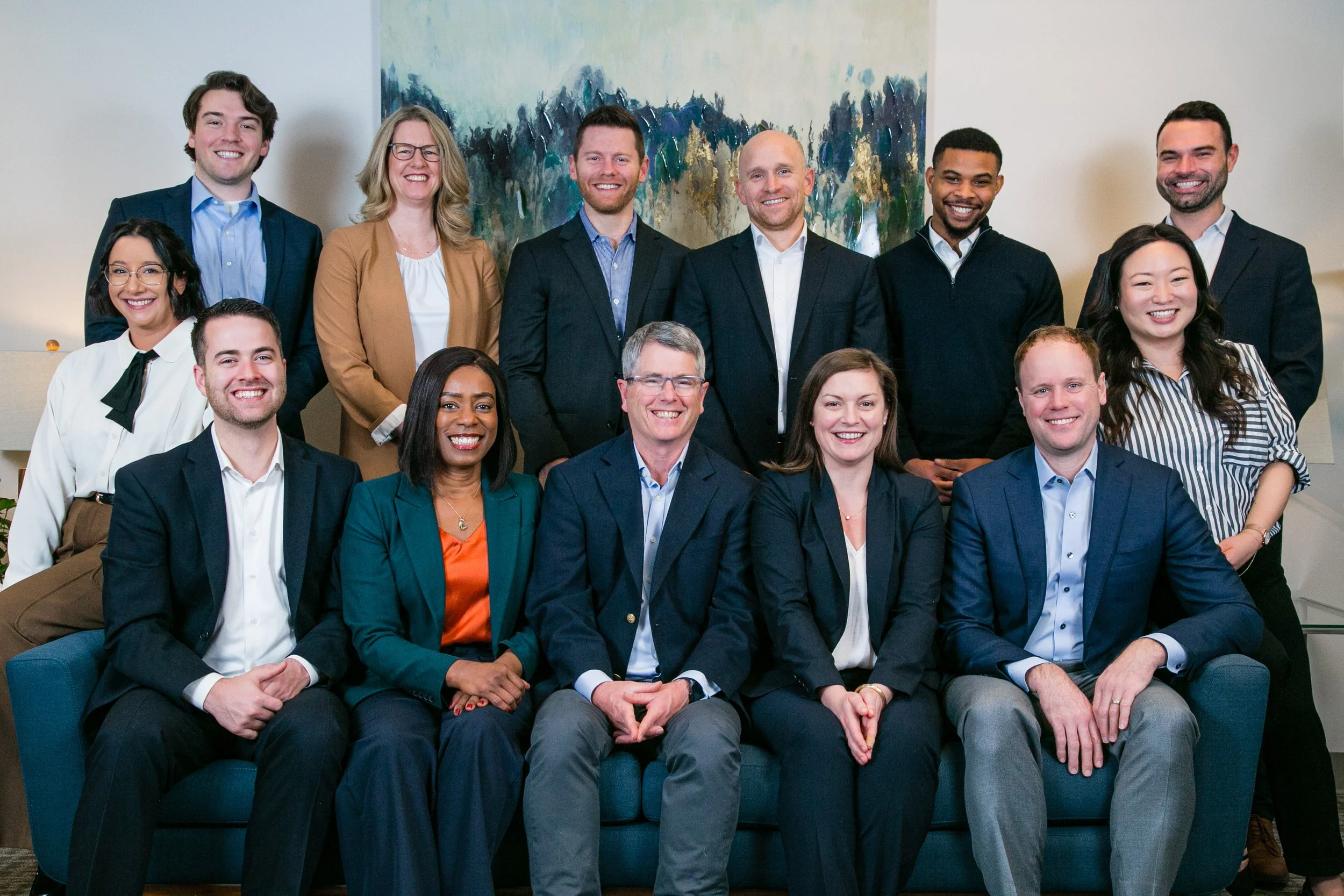 A diverse group of fifteen professionals in business attire posing together in an office setting, with a large abstract painting in the background.