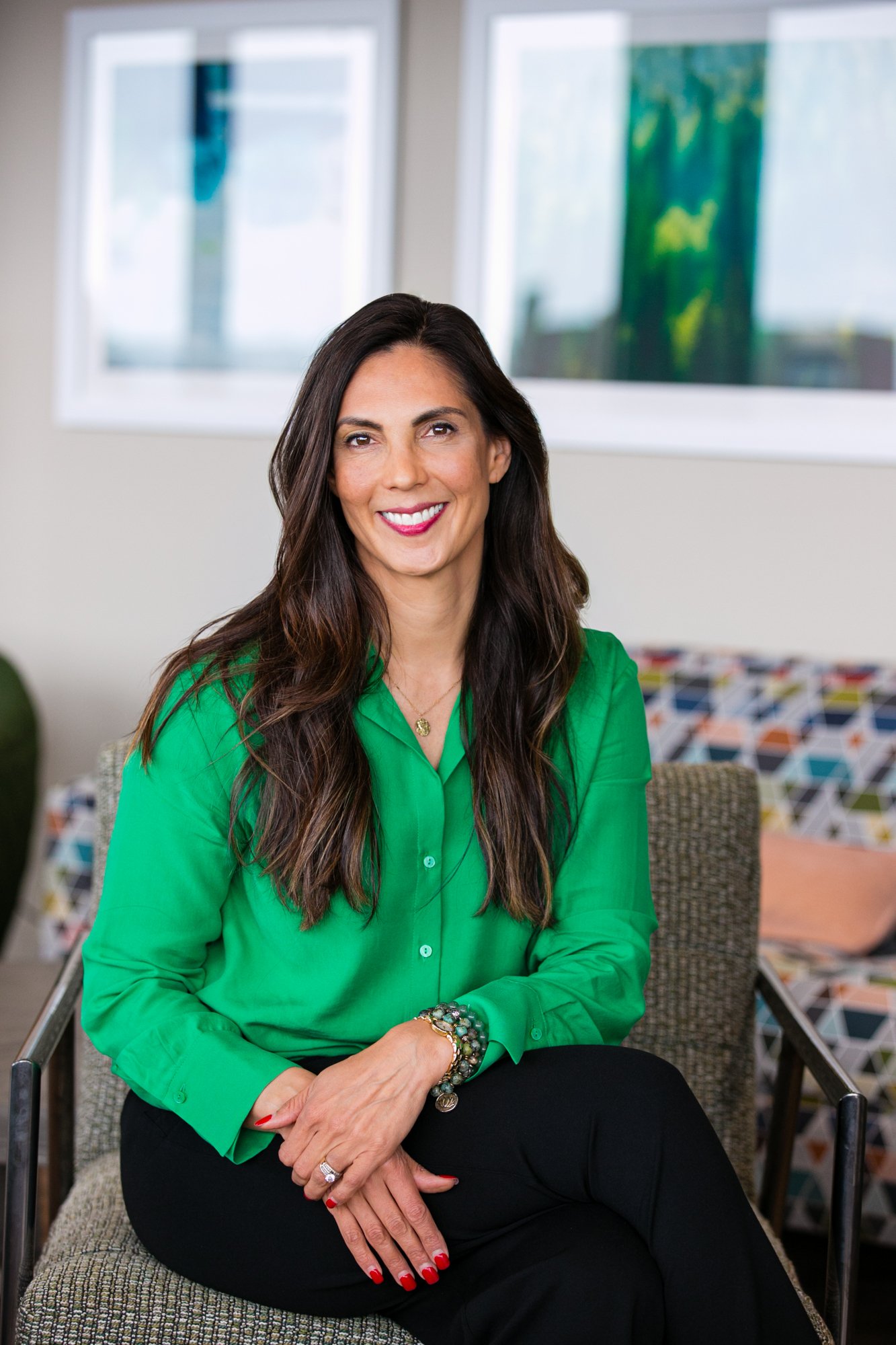 A smiling woman with long brown hair sitting on a patterned chair indoors, wearing a bright green blouse and black pants, with colorful cushions in the background.