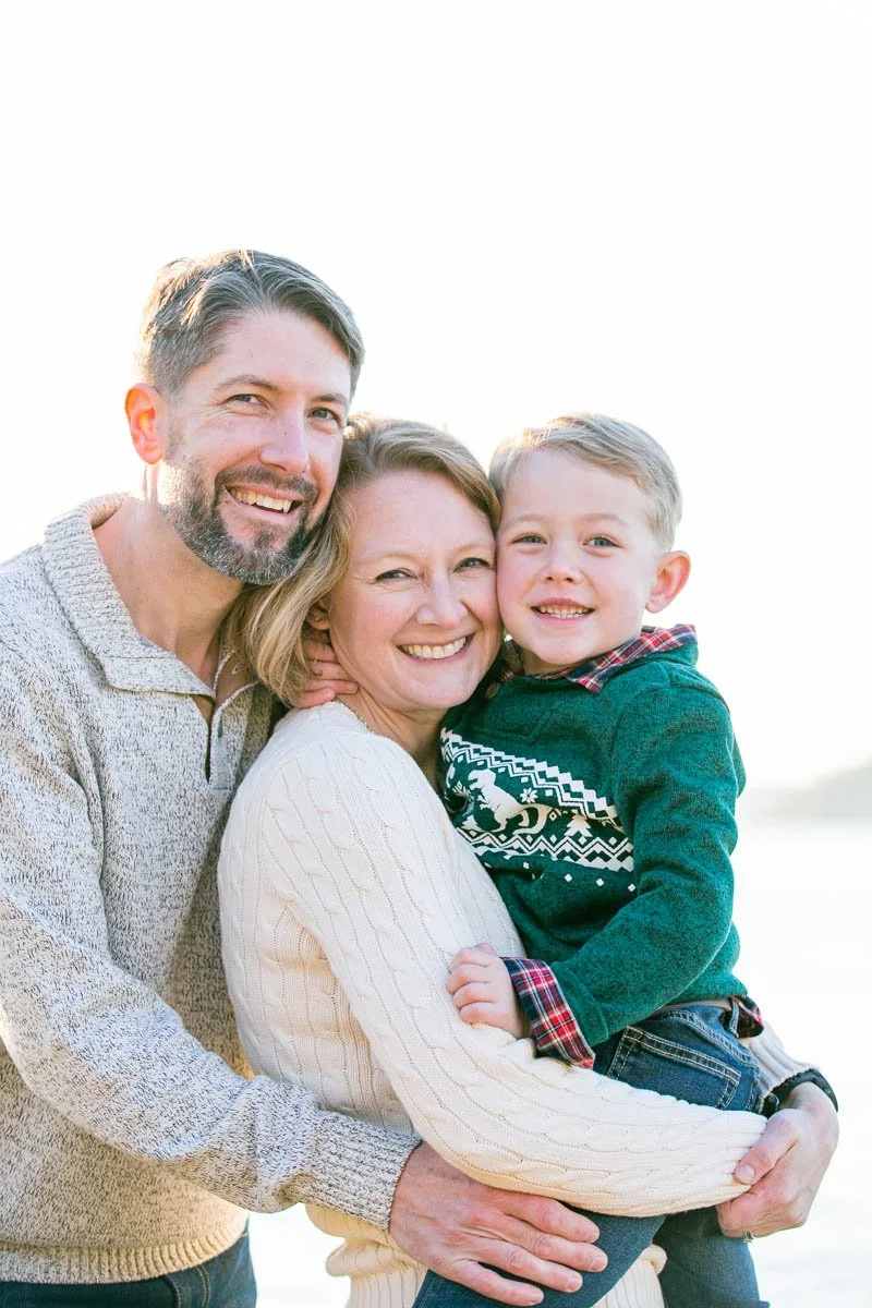 A smiling family of three, a man, woman, and young boy, standing outdoors in bright sunlight.