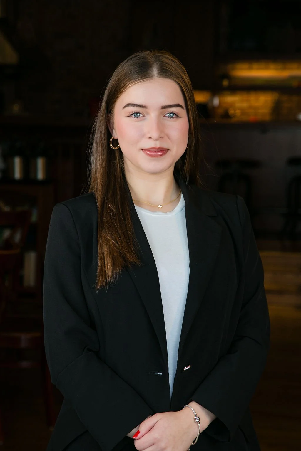 A young woman with long brown hair, wearing a black blazer over a white t-shirt, standing indoors with a dark background. She has light skin, blue eyes, and is smiling softly.