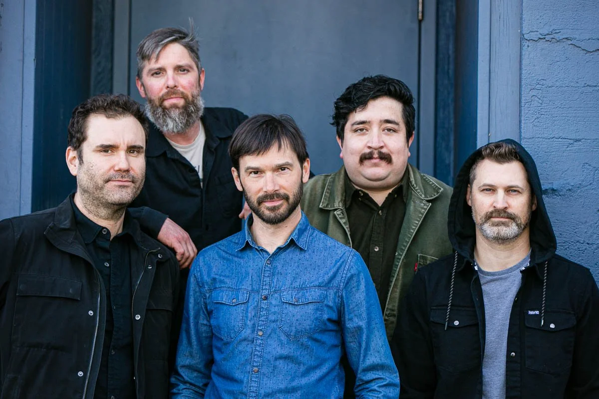 Six men standing together outdoors in front of a blue wall, posing for a photo. They have various facial hair styles and are dressed casually in jackets and shirts.