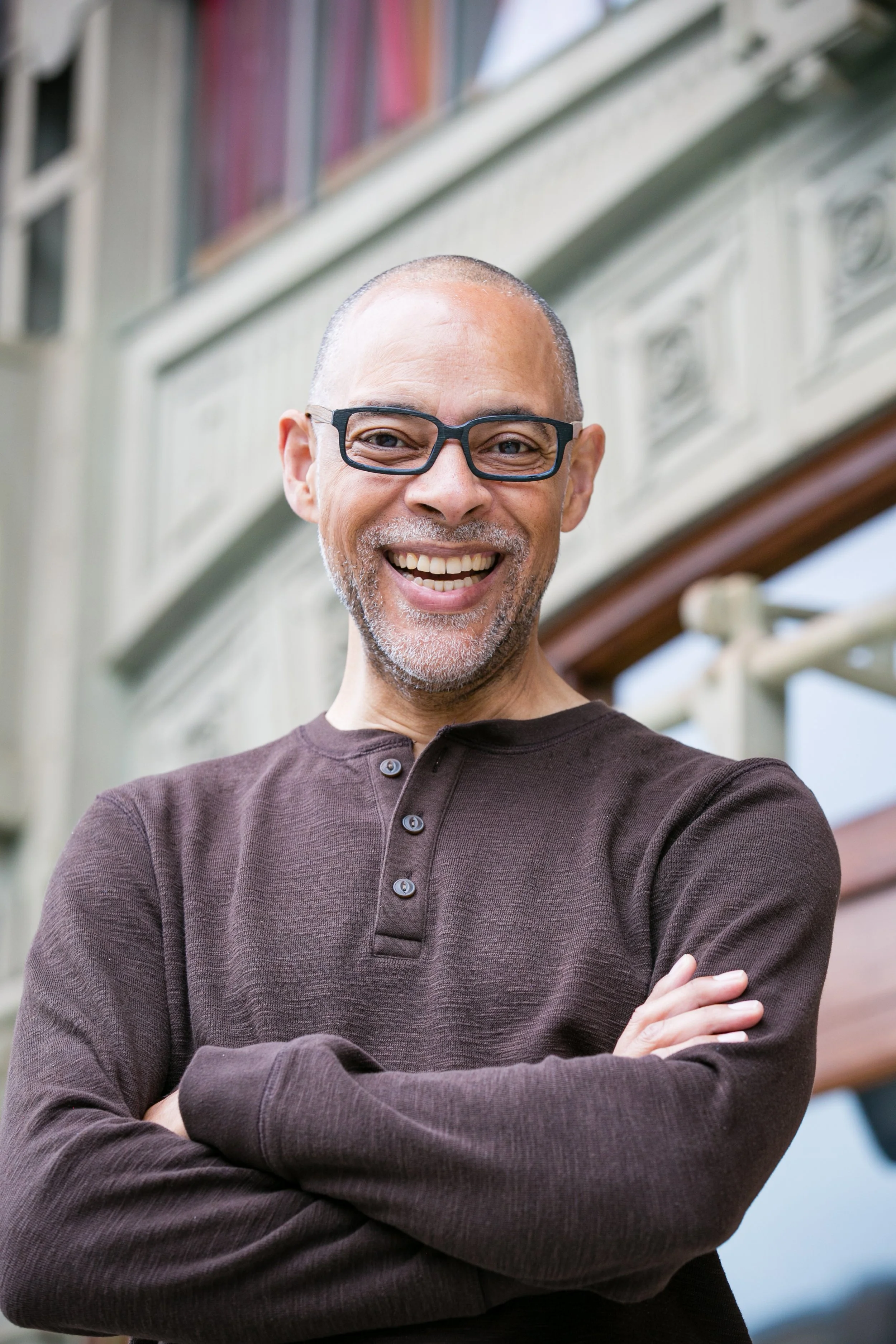 A smiling middle-aged man with glasses and a beard, wearing a brown shirt, standing outdoors with arms crossed in front of a building with bookshelves visible through a window.