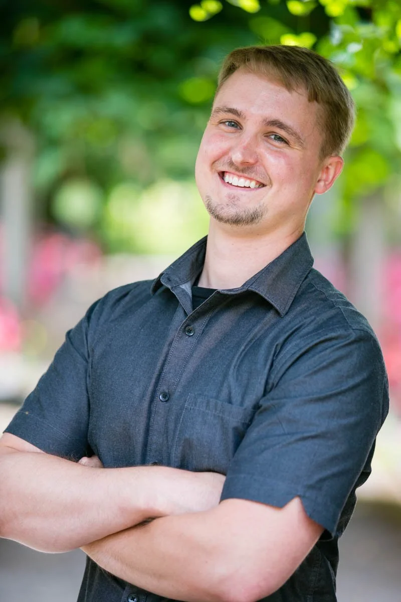 A young man smiling with arms crossed, standing outdoors with green trees and pink flowers in the background.