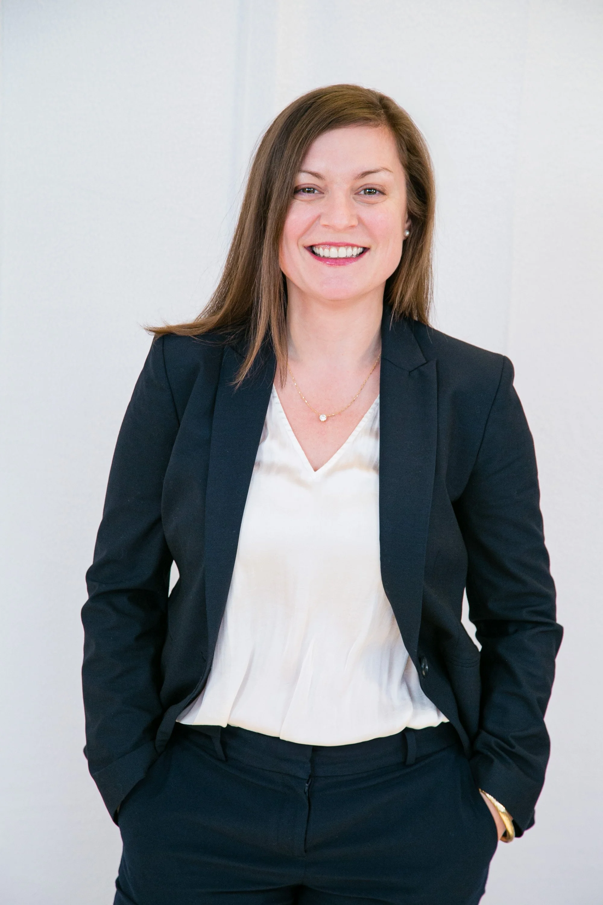 A woman with brown hair smiling, dressed in a black blazer, white blouse, and black pants, standing against a plain background.