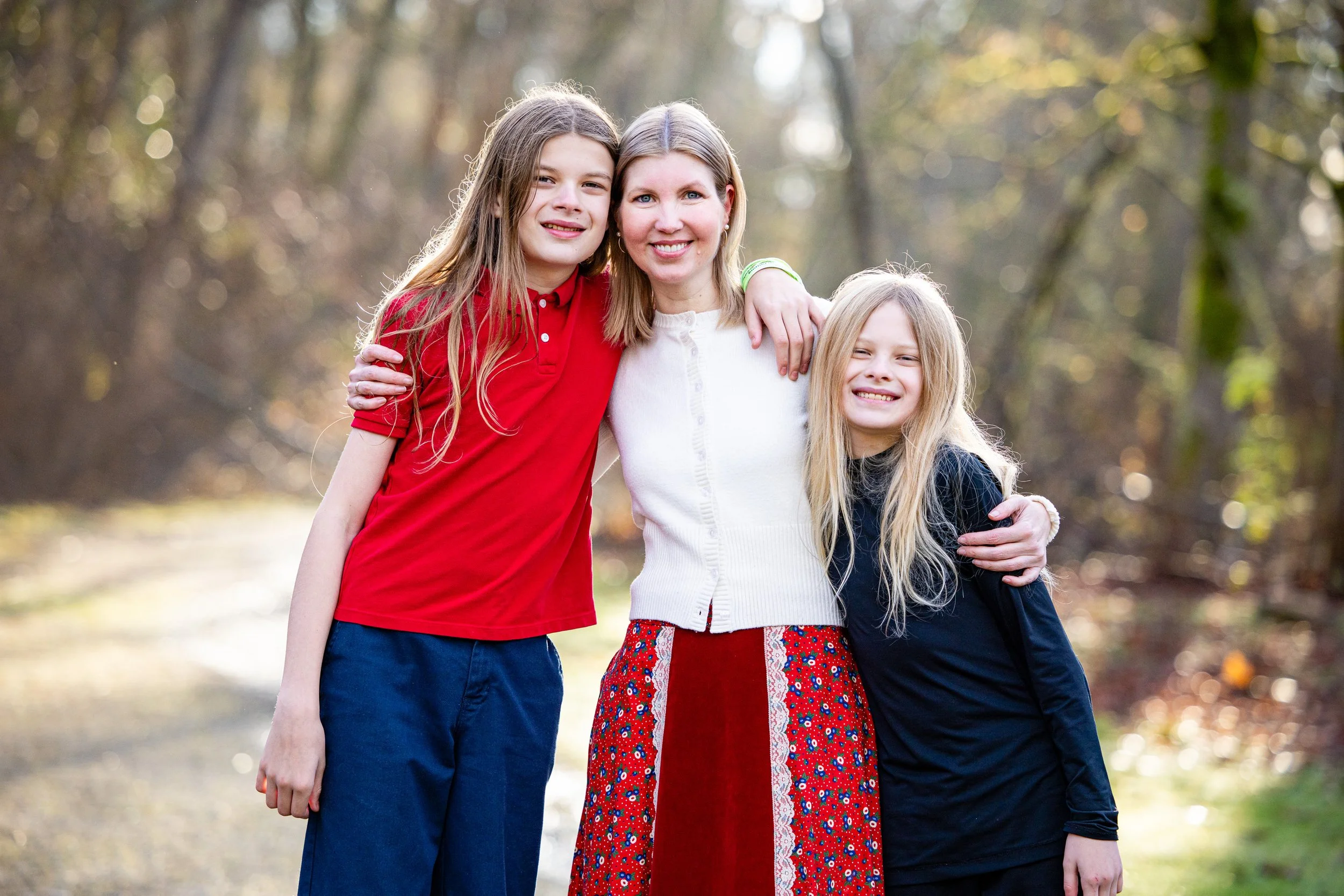 A woman and two girls smiling outdoors in a wooded area, with the women and girls embracing each other.