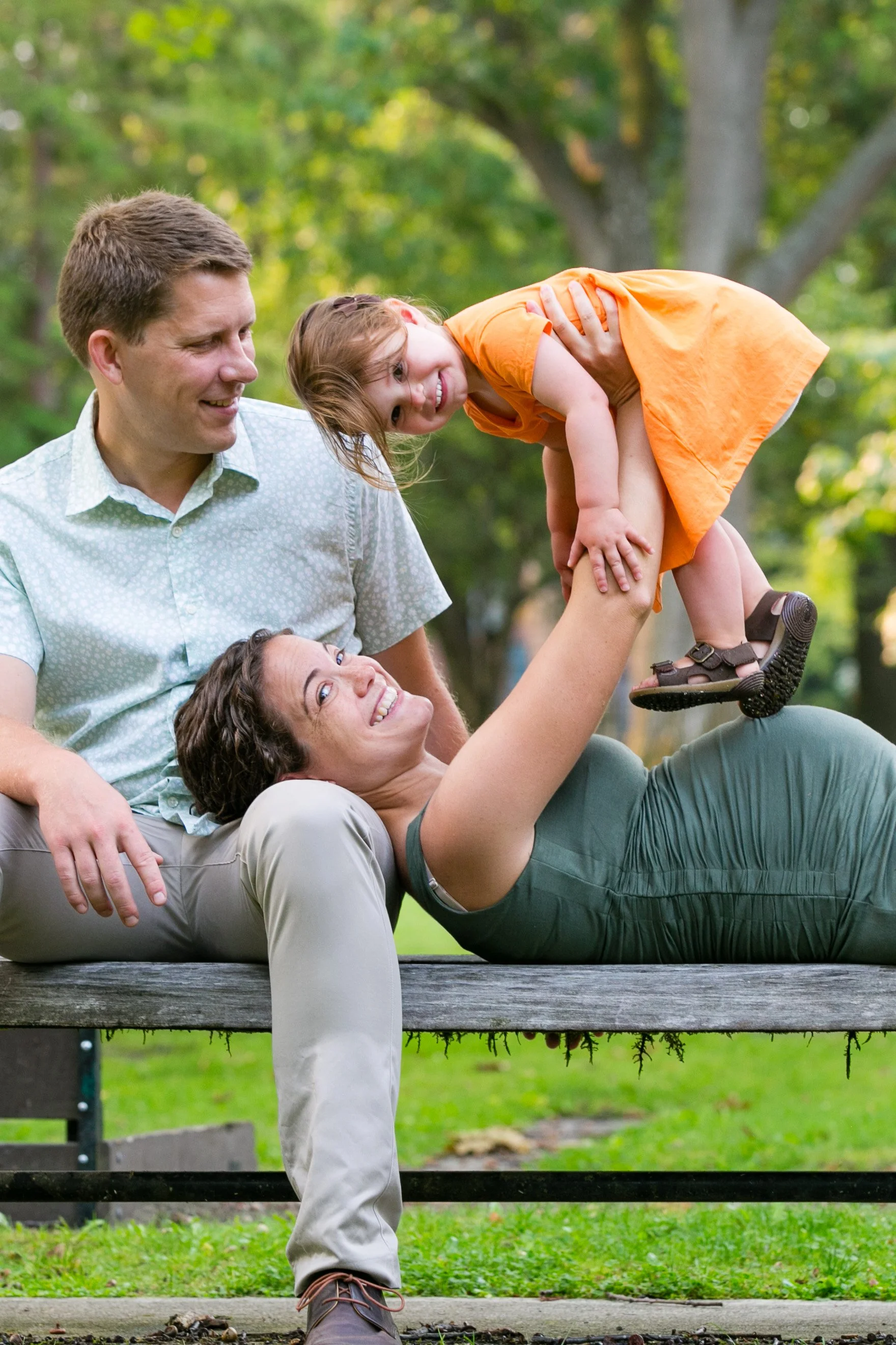 Family of four enjoying outdoor play on a park bench, with a woman lying down, a man sitting beside her, and a young girl in an orange dress bouncing on her mother's legs.