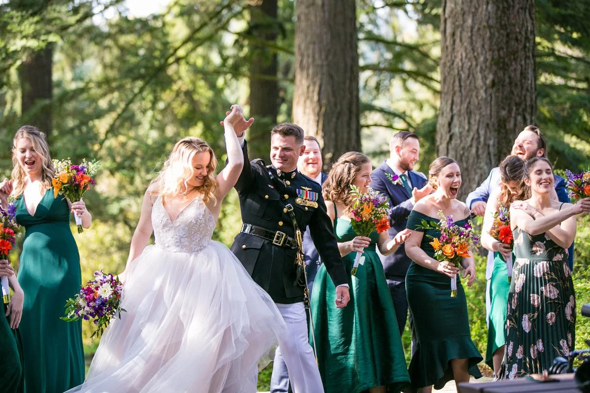 A bride and a groom in a military uniform are walking together and holding hands in a forest. They are surrounded by friends in formal dresses and suits, who are celebrating, smiling, and holding colorful bouquets of flowers.