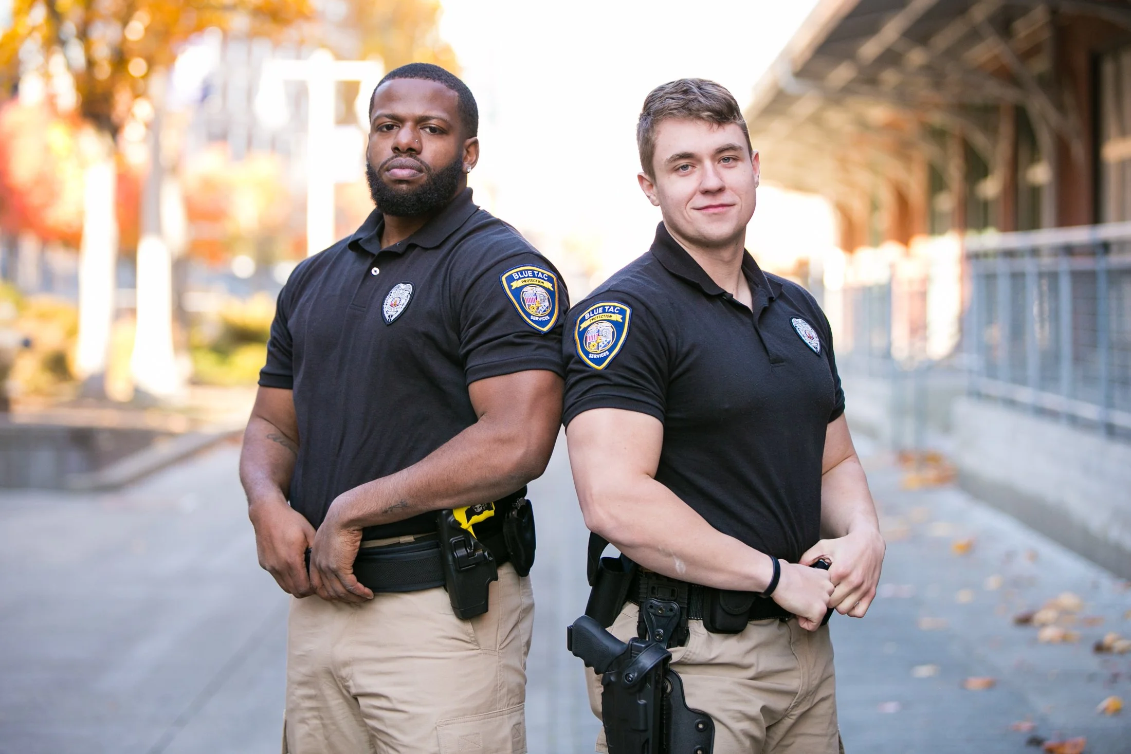 Two police officers standing outdoors on a sidewalk, side by side, looking at the camera, with trees and a building in the background.