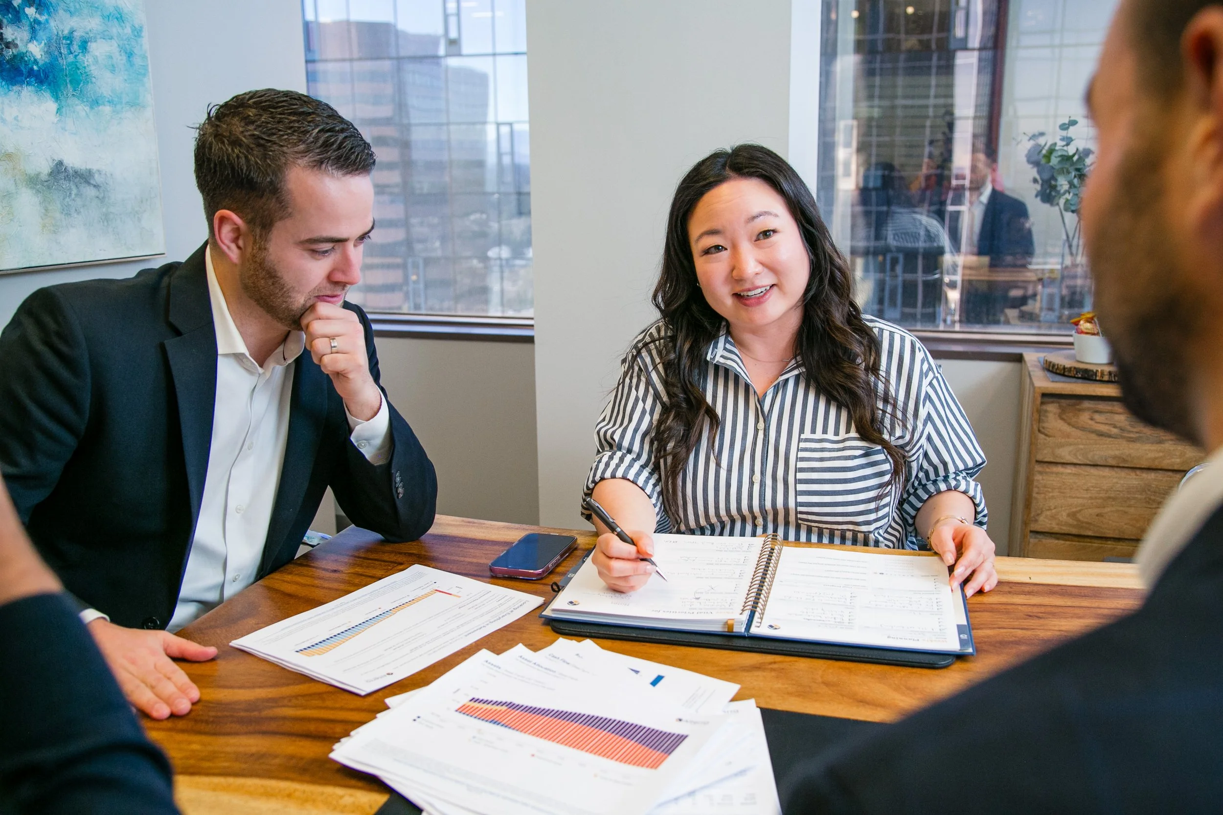 People in a business meeting at a conference table with documents, a phone, and a notebook, in an office with large windows.