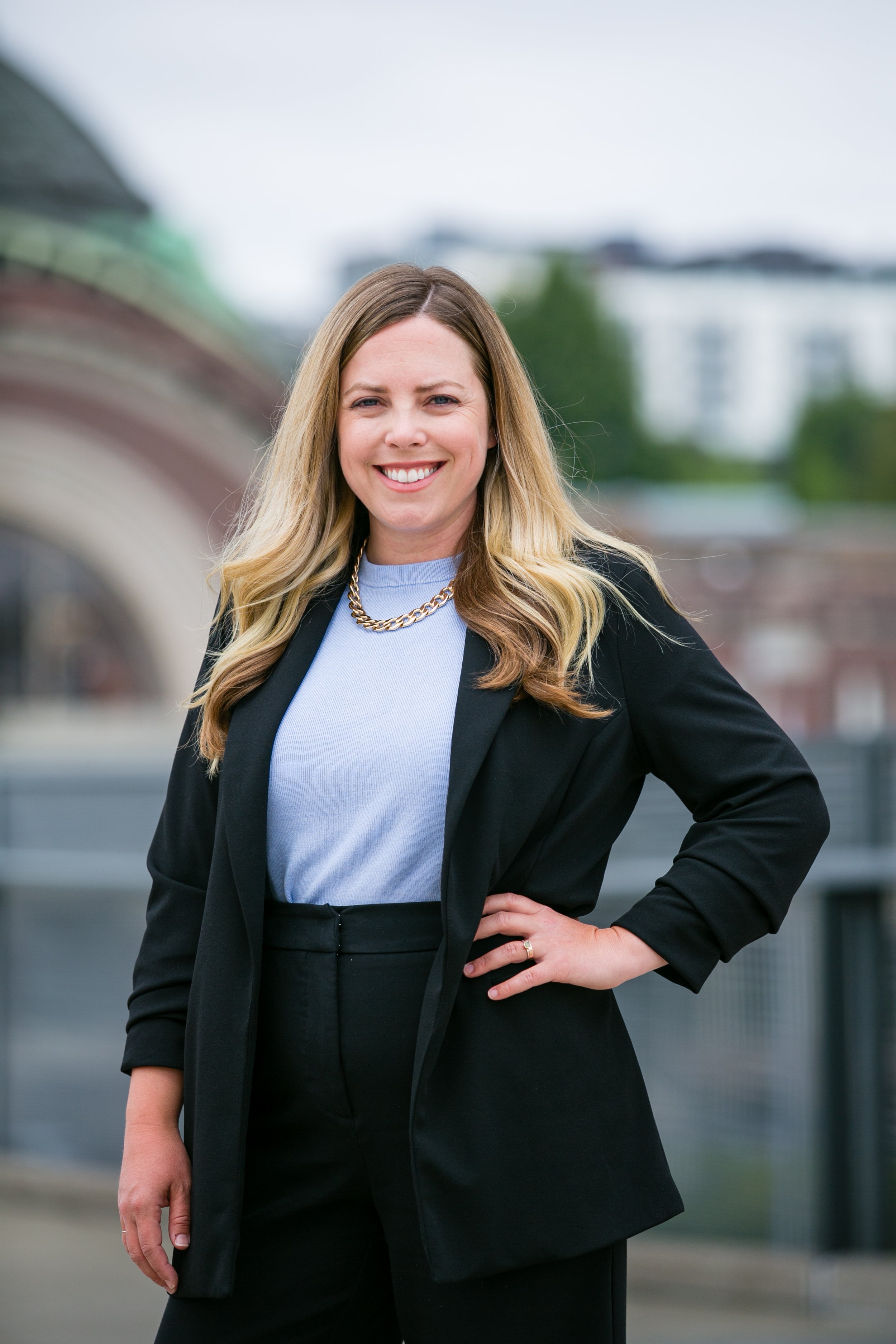 A smiling woman with blonde hair in a black blazer, white blouse, and gold chain necklace, standing outdoors with a cityscape in the background.