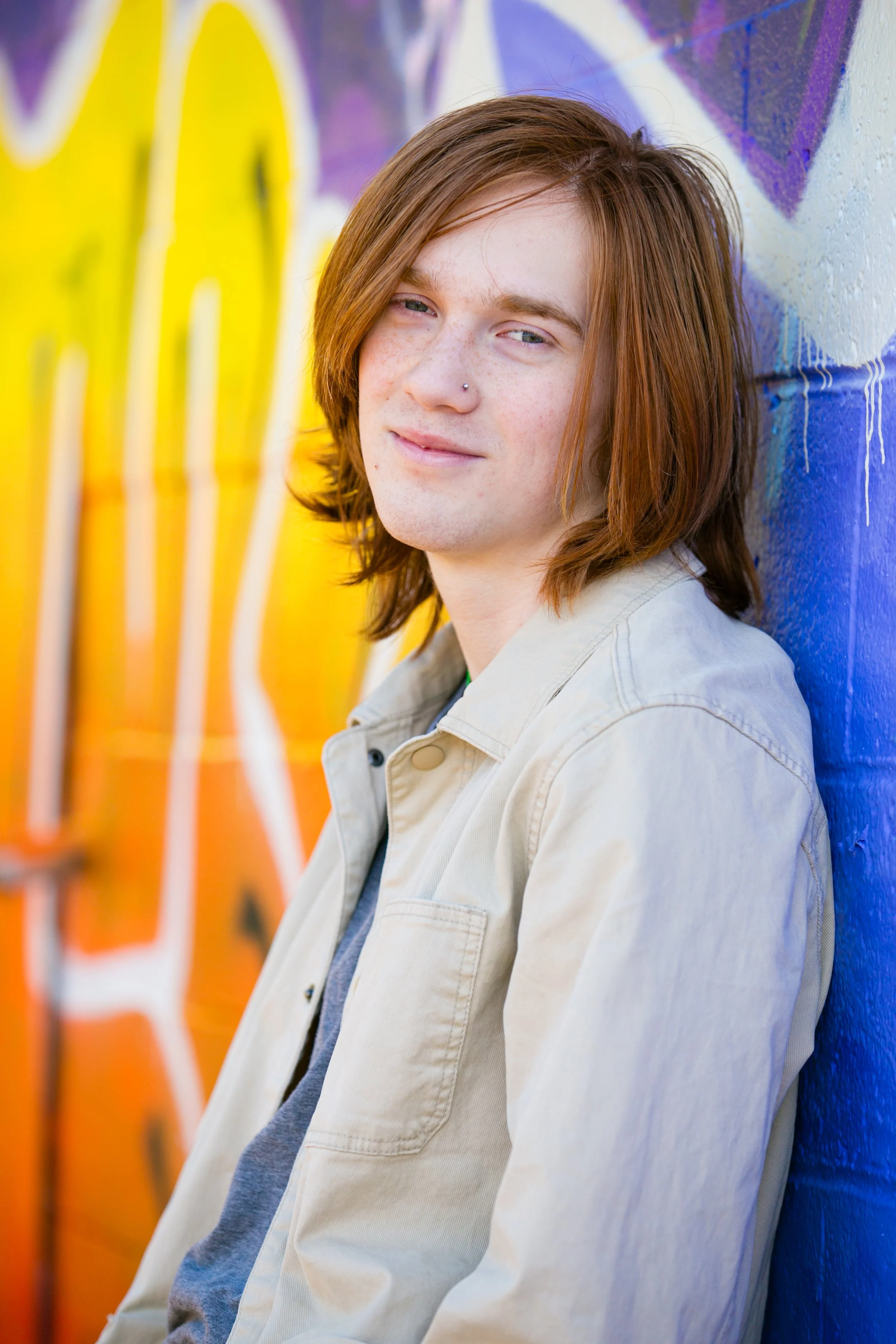 A young man with shoulder-length red hair, wearing a light beige jacket and gray shirt, standing against a colorful, graffiti-covered wall.