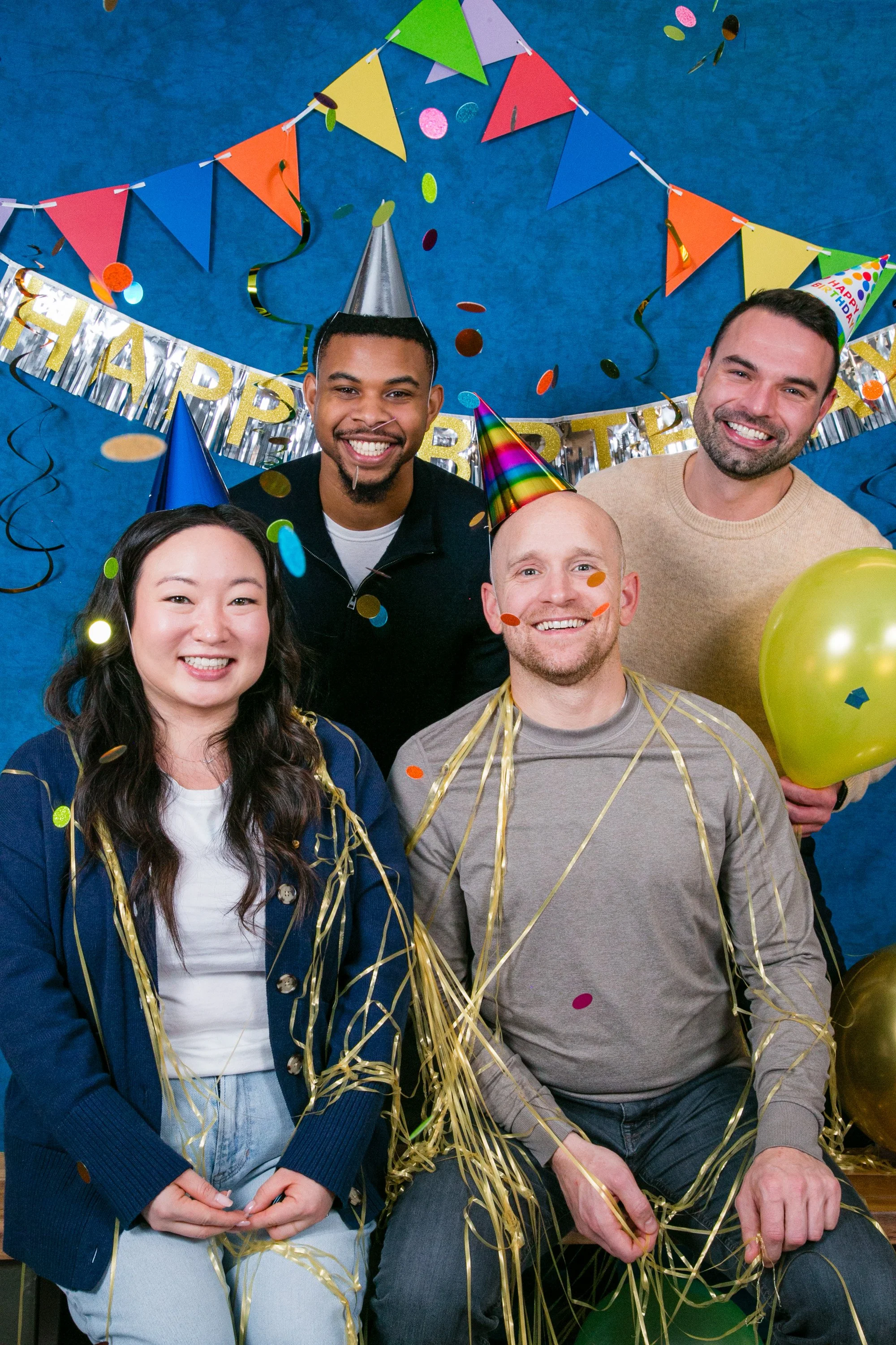 Group of five people celebrating a birthday with colorful party hats, balloons, confetti, and a decorative banner that reads "HAPPY BIRTHDAY," against a blue background.