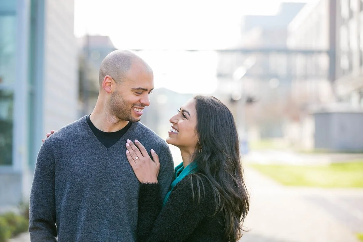 A smiling man and woman looking at each other outside in a city environment, with the woman touching the man's chest.