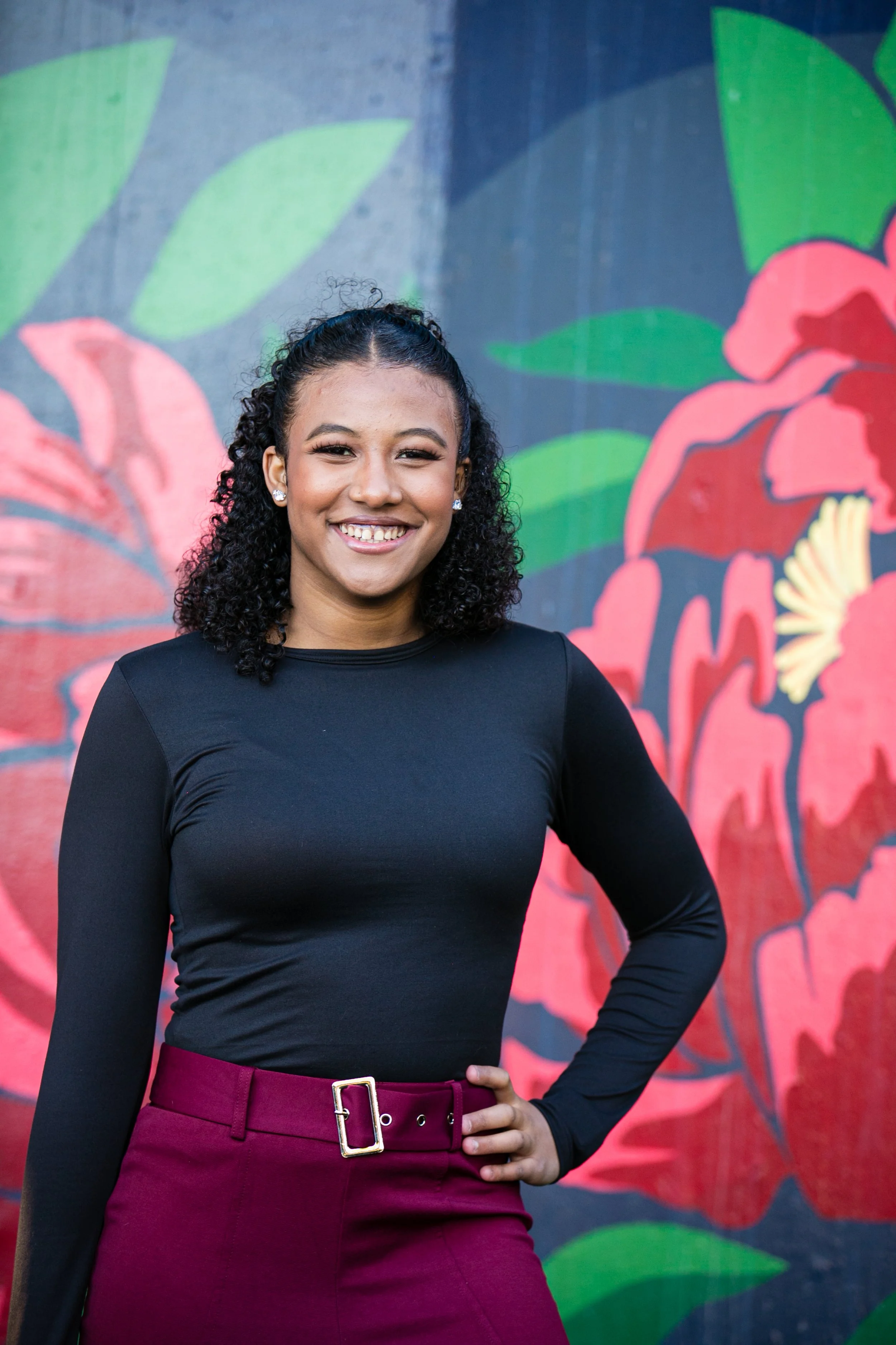 A smiling woman with curly black hair, wearing a black long-sleeve top and burgundy pants with a gold buckle belt, standing in front of a colorful floral mural.