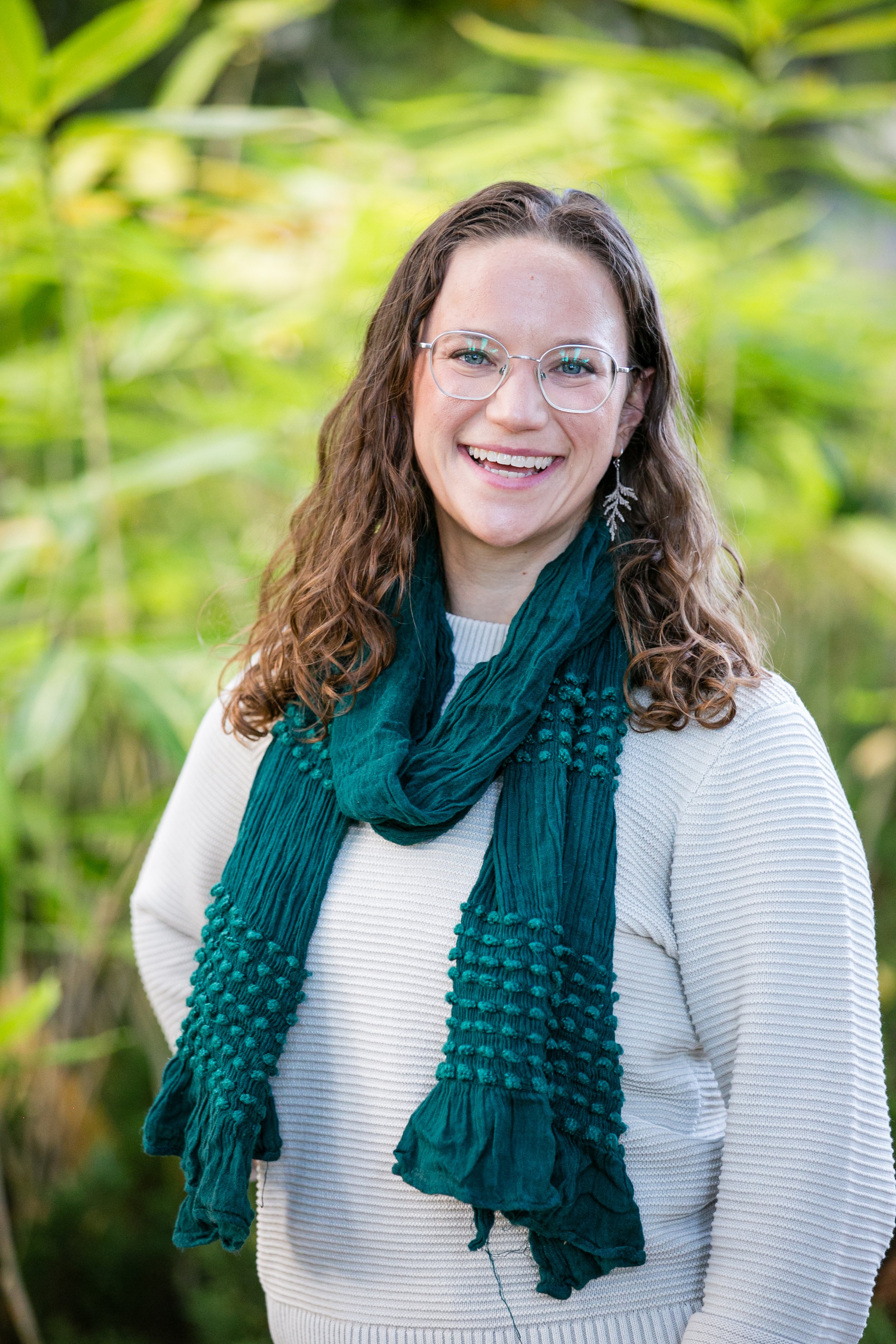 A smiling woman with curly brown hair, glasses, and dangling earrings stands outdoors in a green, leafy background. She wears a white textured sweater and a teal scarf.