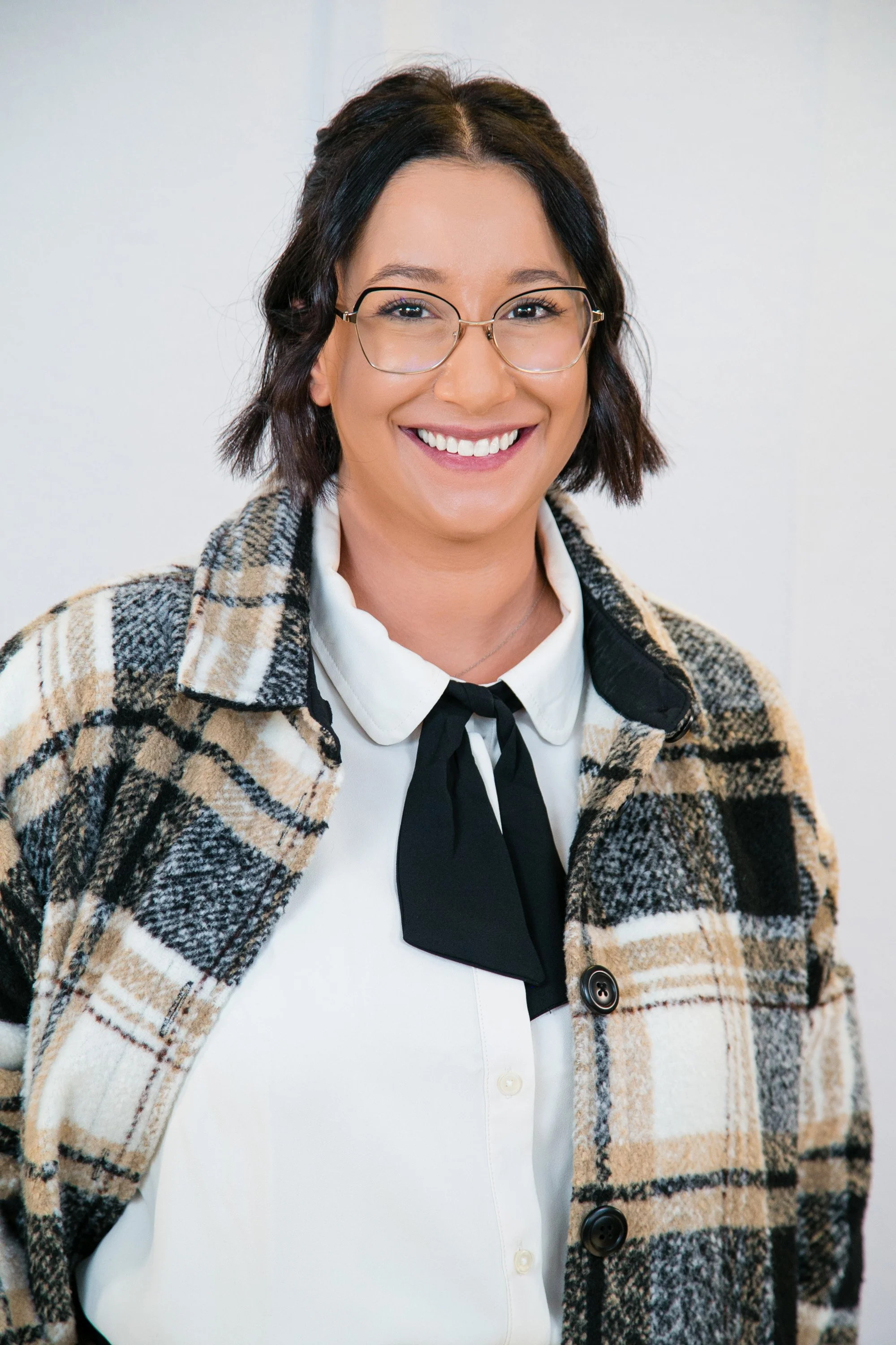 A woman with short dark hair, glasses, and a bright smile, wearing a plaid coat over a white blouse with a black bow tie, standing indoors with a white wall background.