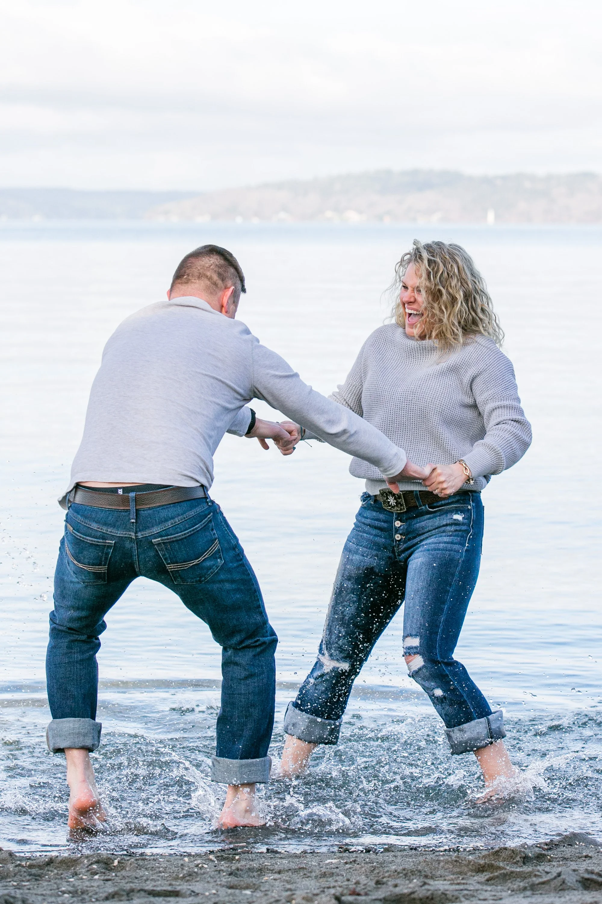 A man and woman playing tug of war in the water at the beach, both smiling and splashing water, with a lake and hills in the background.