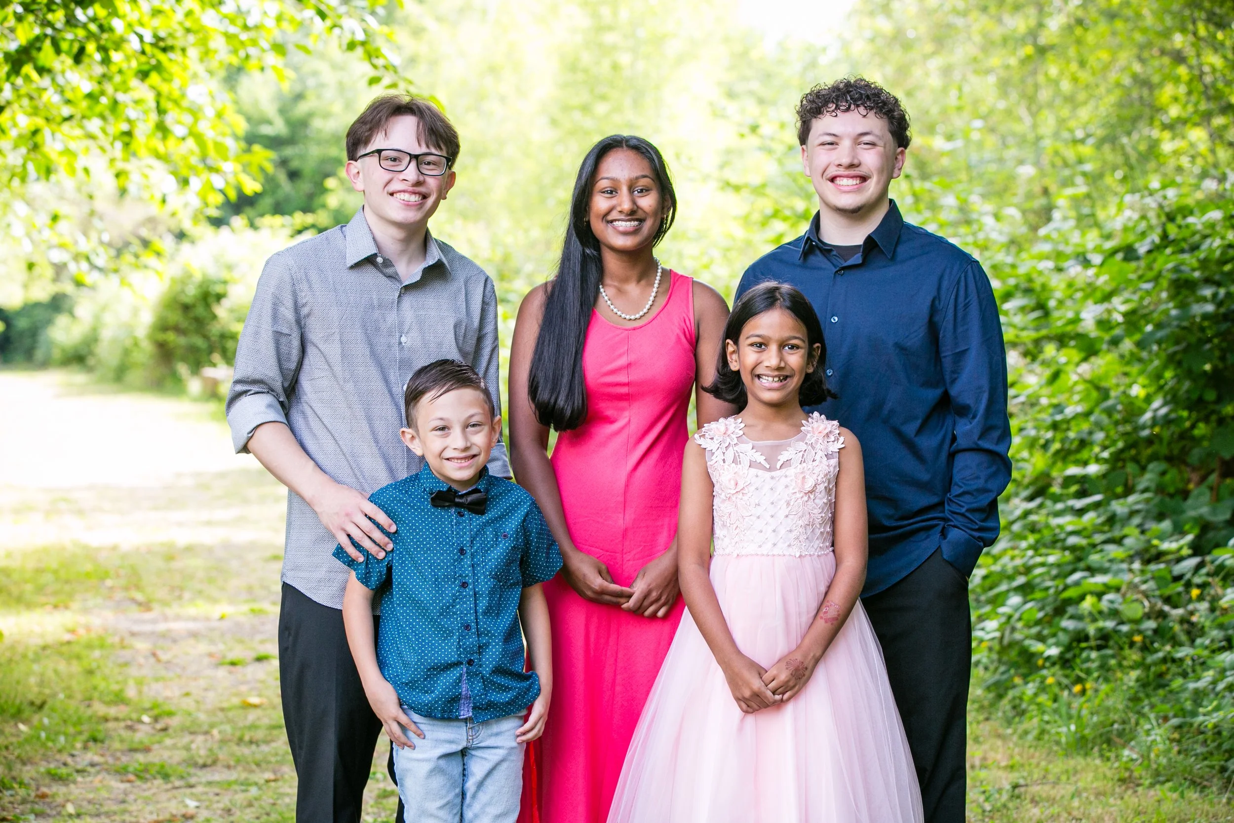 A diverse group of six people standing together outdoors on a sunny day, smiling at the camera. They include three boys and two girls, with a woman in the center. The background features lush green trees and grass.