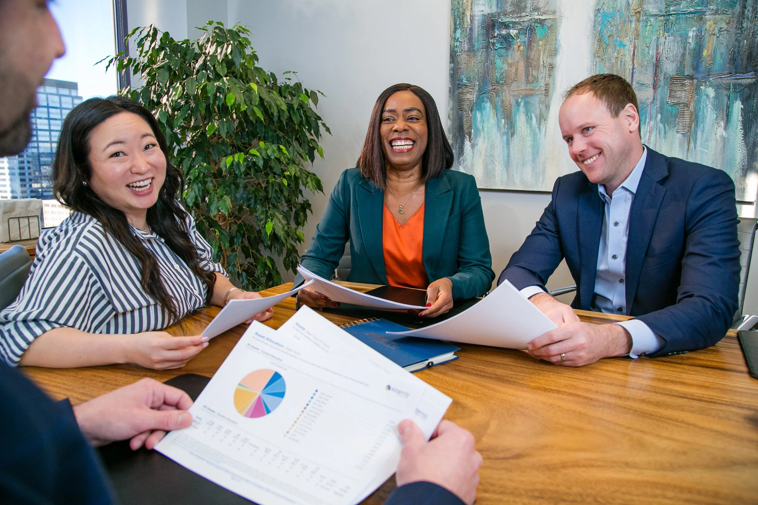 Four business people sitting at a meeting table, talking, and smiling in an office with large windows and a modern painting on the wall.