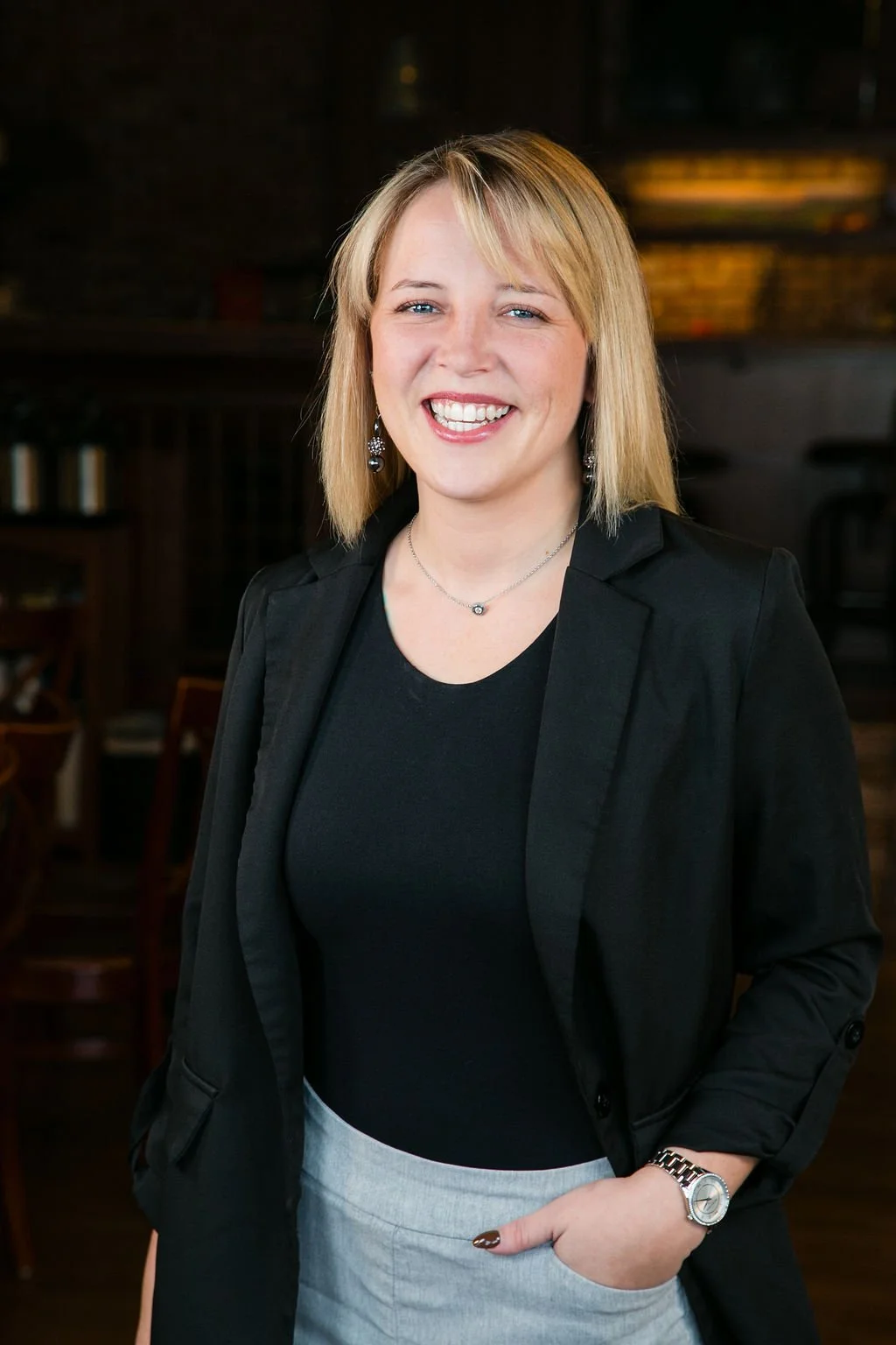 A woman with blonde hair smiling, wearing a black blazer, a black top, a silver necklace, and a silver watch, standing indoors in a dimly lit room with wooden furniture and bookshelves in the background.