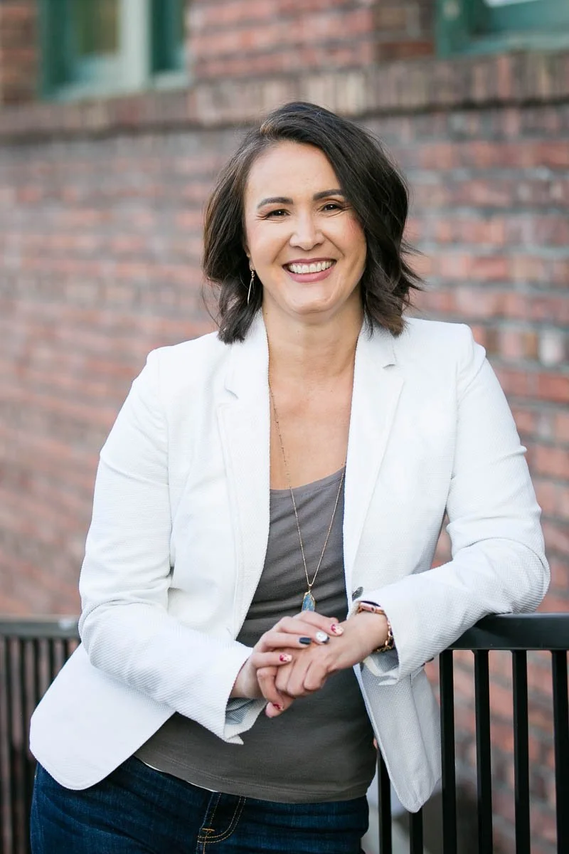 Woman with dark hair smiling, dressed in a white blazer and gray top, standing outdoors against a brick wall.